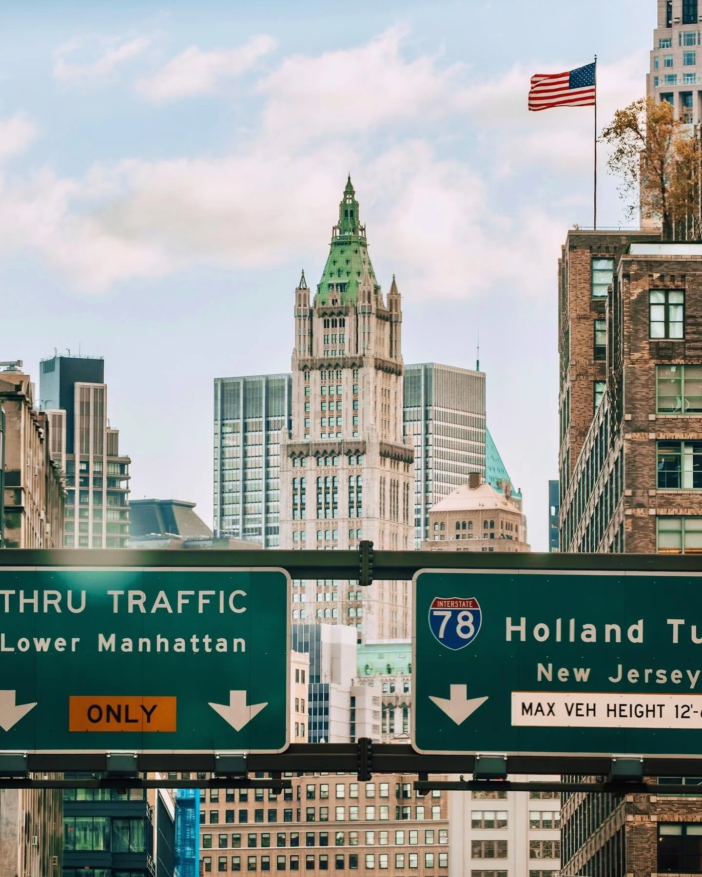 New York City street scene showing tall buildings, including a historic skyscraper with a green roof, and a green highway sign indicating traffic directions toward Lower Manhattan and New Jersey with American flags in the background.