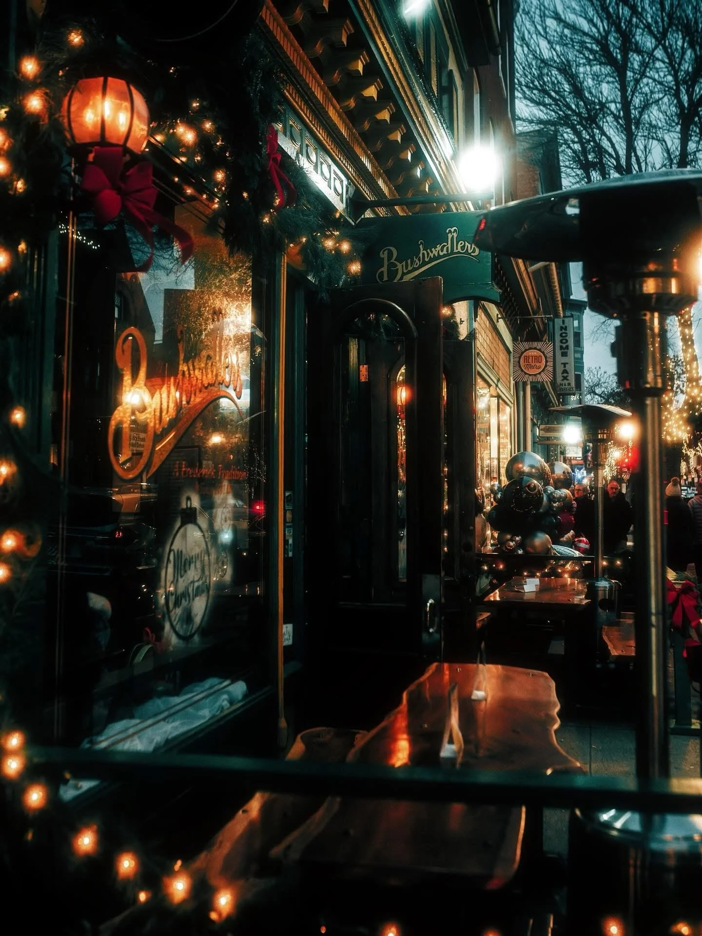 A decorated storefront with Christmas lights, wreaths, and ornaments, with people walking outside on a city street during evening.