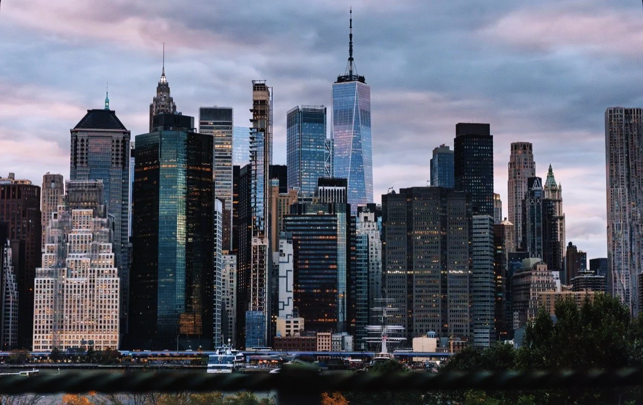 Skyline of New York City with skyscrapers, including One World Trade Center, under a cloudy sky at dusk.
