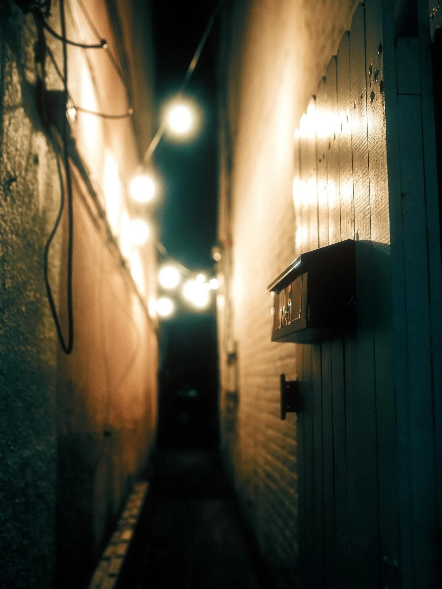 A narrow alleyway illuminated by string lights at night, with a wall-mounted mailbox in the foreground.
