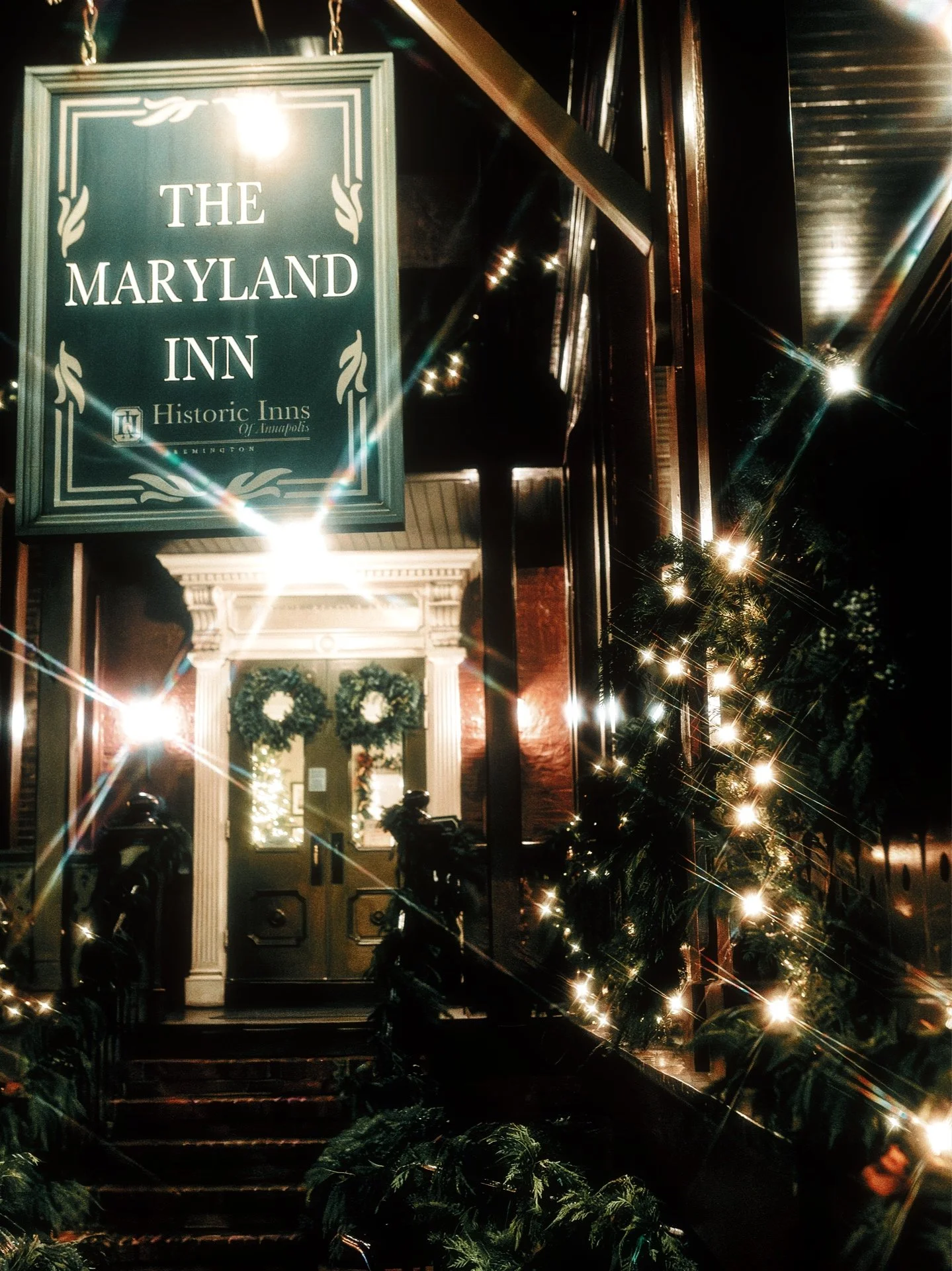 Nighttime view of the entrance to The Maryland Inn, a historic inn in Indianapolis, decorated with Christmas wreaths and lights, with a sign overhead reading 'The Maryland Inn Historic Inns of Indianapolis'.
