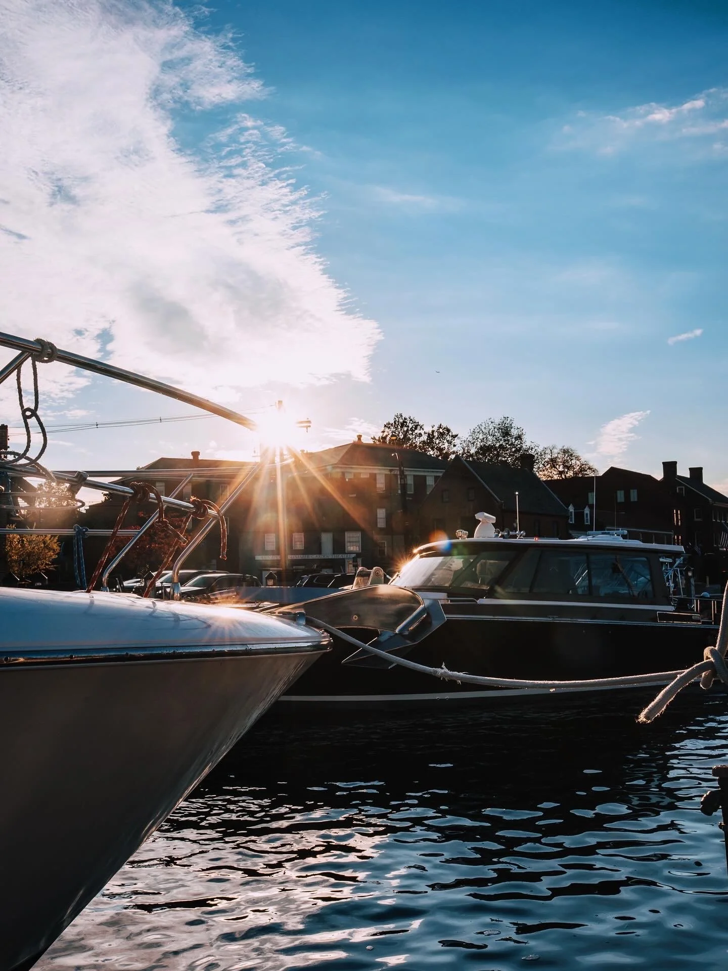 A marina at sunset featuring boats docked on calm water, with a clear sky and buildings in the background.
