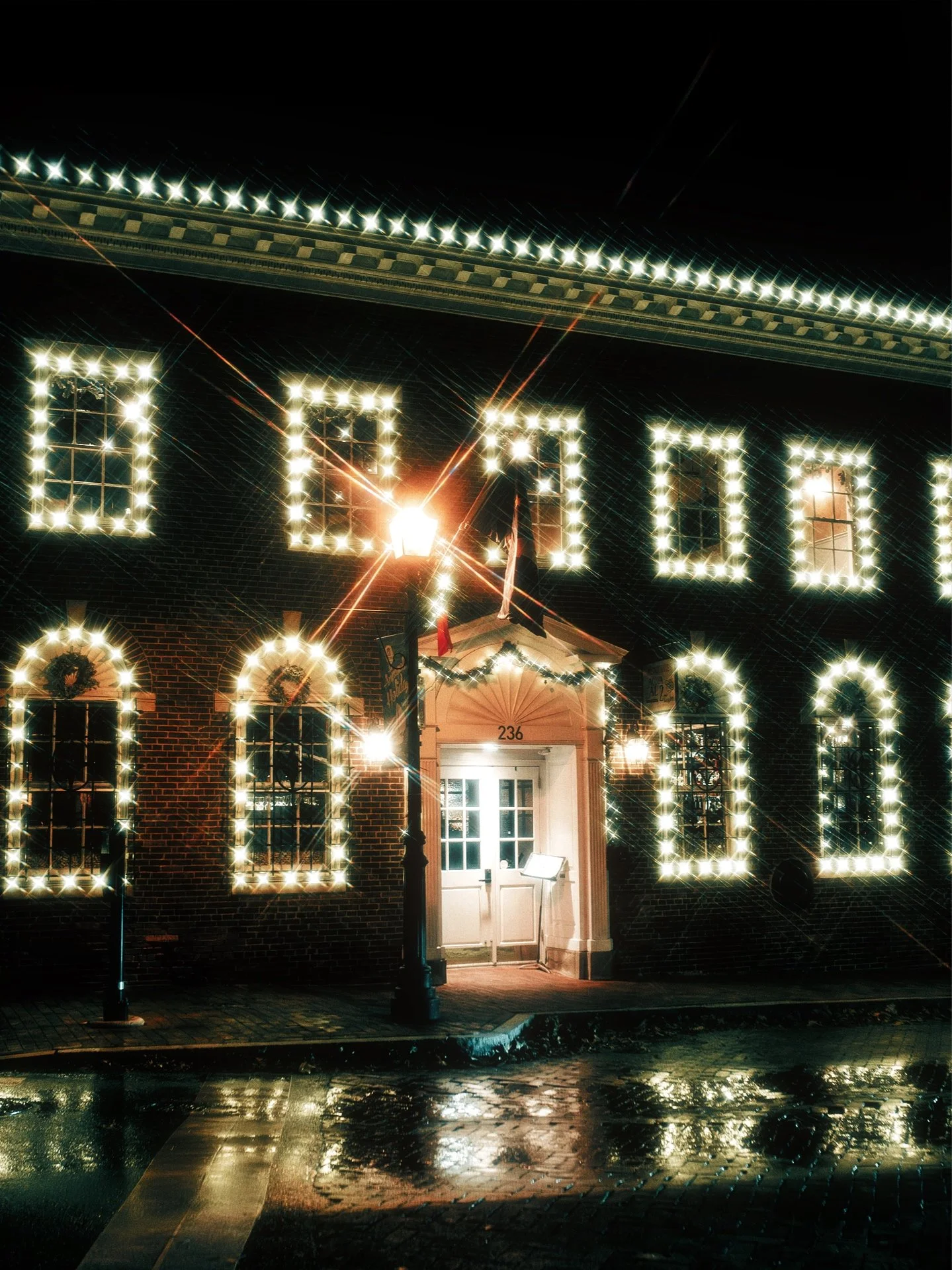 A brick building decorated with bright Christmas lights around windows and entrance at night, with wet pavement reflecting the lights.