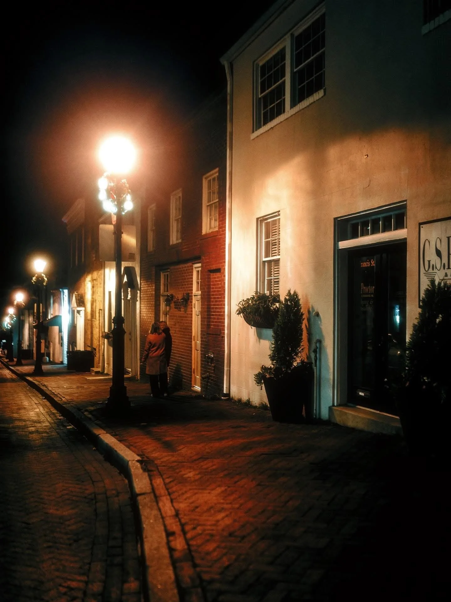 A quiet city street at night with street lamps illuminating the sidewalk, storefronts, and a couple walking together.
