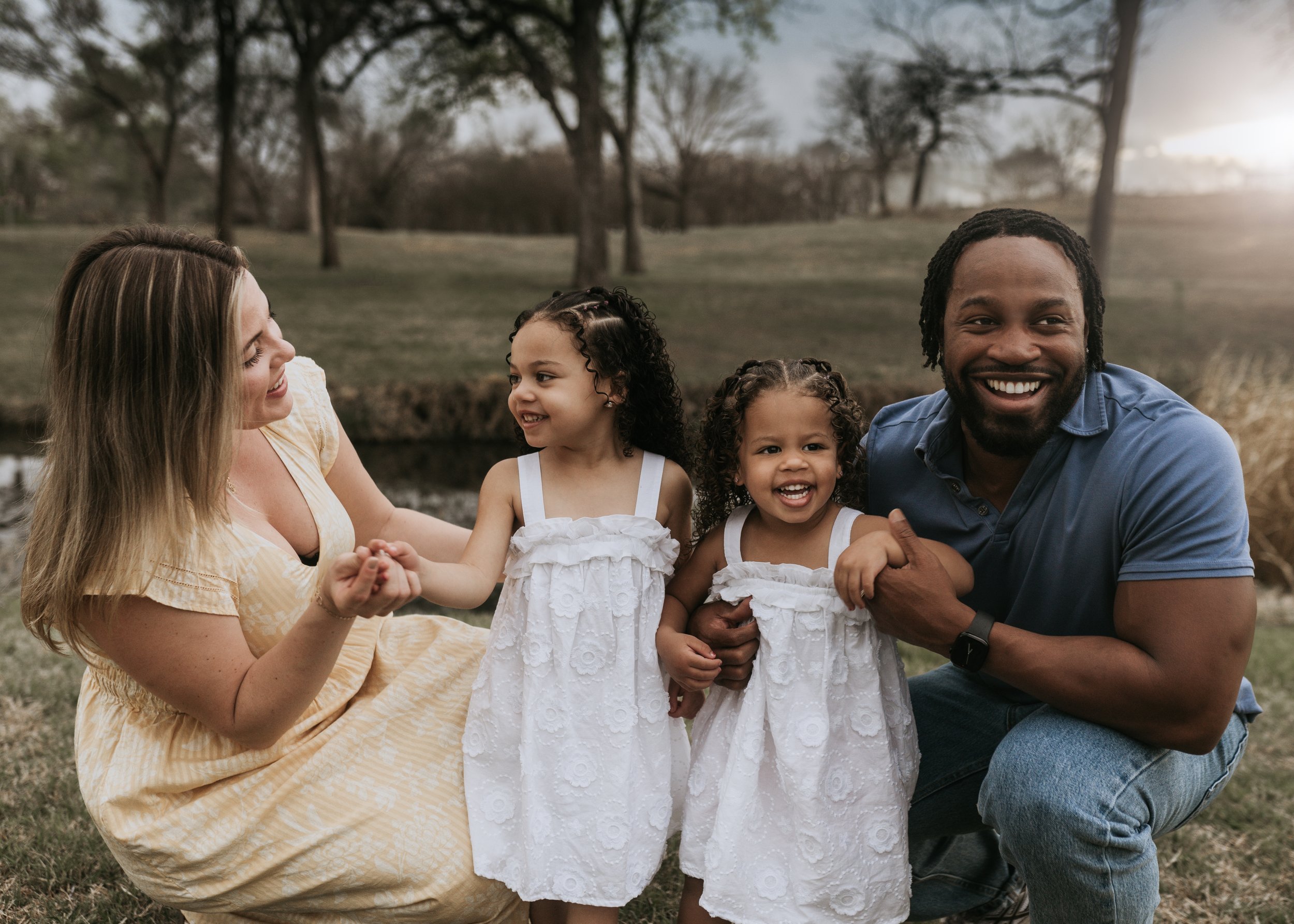 A family of four smiling and posing outdoors in a park. The mother and father are kneeling, holding hands with their two young daughters, who are wearing matching white dresses. The setting is grassy with trees in the background.