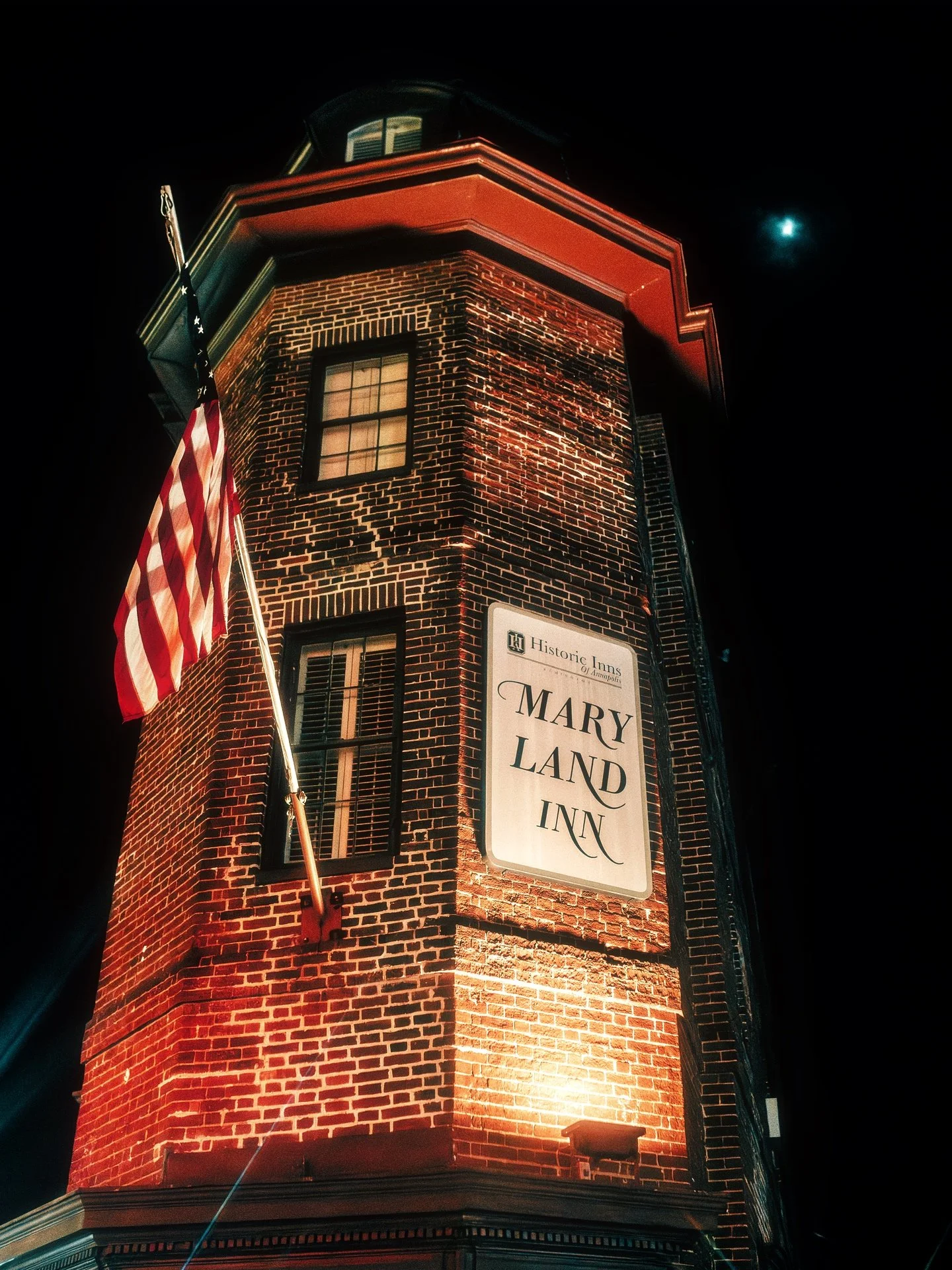 Nighttime photo of a brick building with a sign that reads 'Mary Land Inn' and an American flag hanging from a pole. The building has windows and is illuminated by lights, with a visible moon in the dark sky.