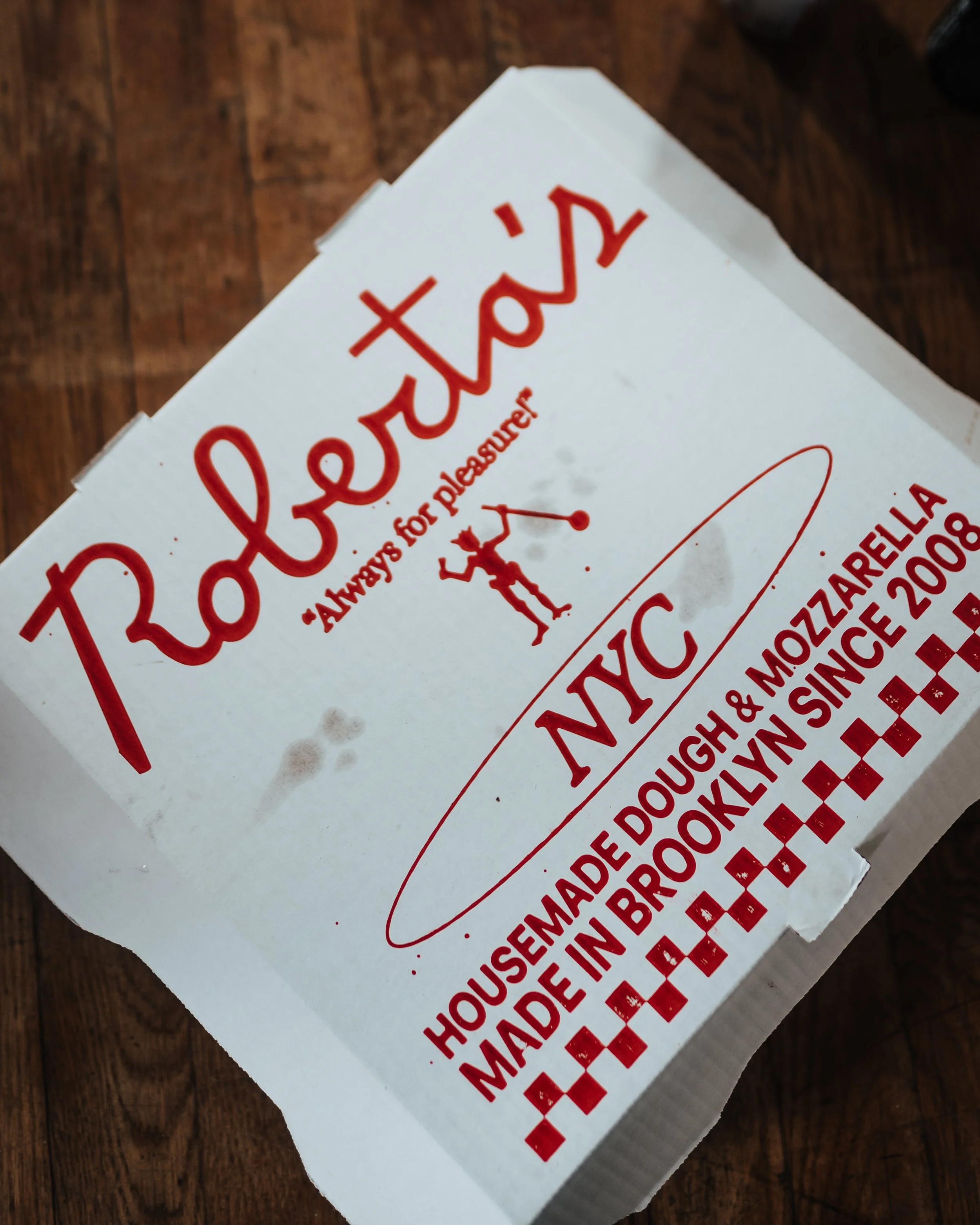 Close-up of a Pizza Hut pizza box with red and white branding, depicting the company's logo, a small elf character, and text indicating the pizza is made in Brooklyn since 2008.