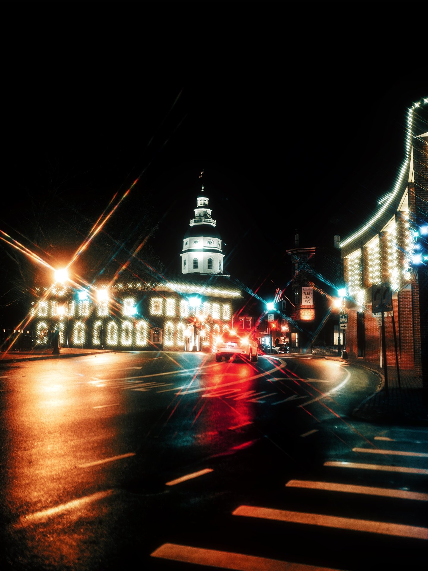 Nighttime scene of a historic church with a tall steeple and surrounding buildings lit with bright-colored lights, with blurry street lights and a crosswalk in the foreground.