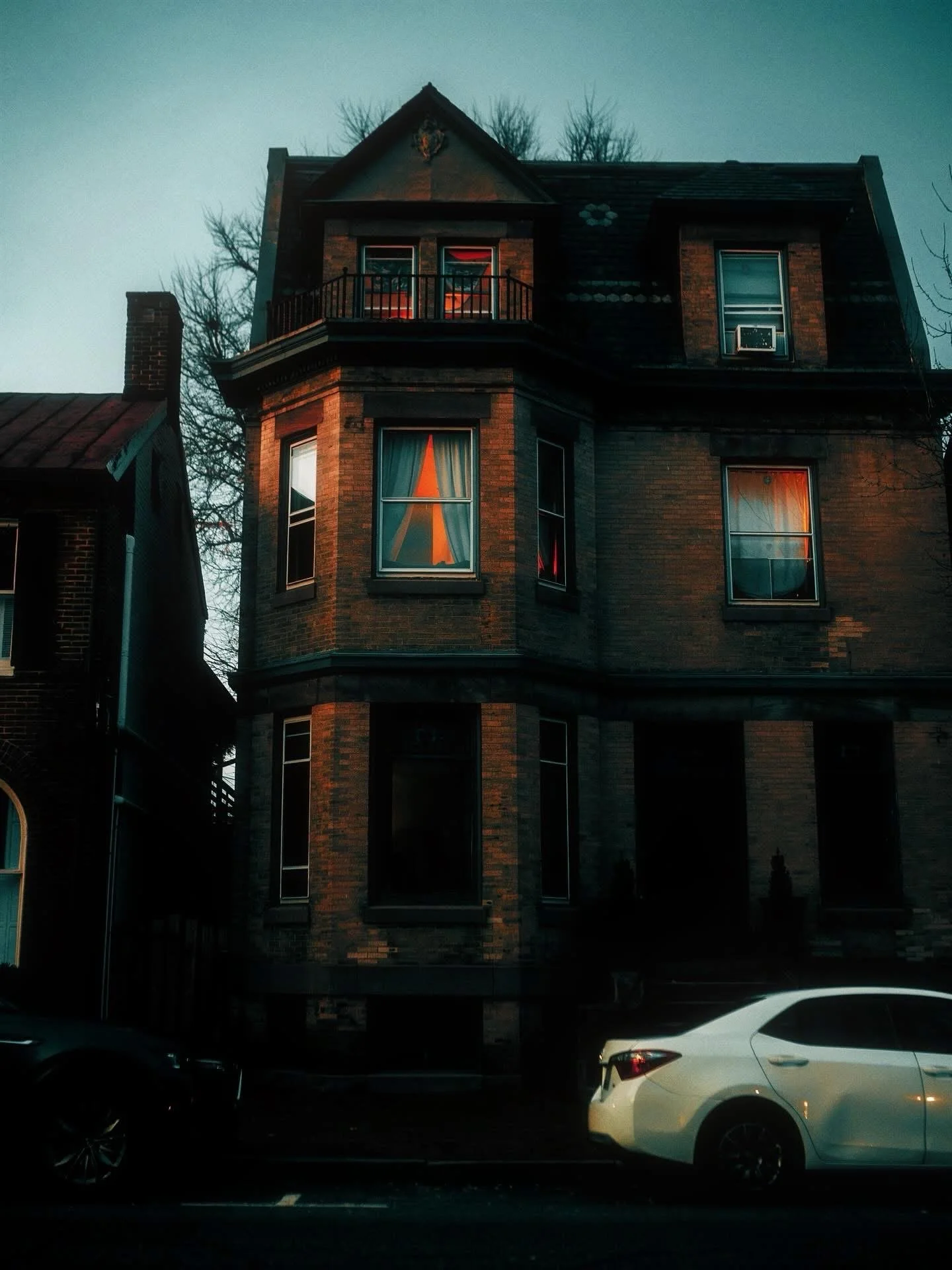A tall, three-story brick house with Victorian-style architecture, featuring a turret with a balcony on the top floor, and windows with curtains. There are cars parked in front of the house.