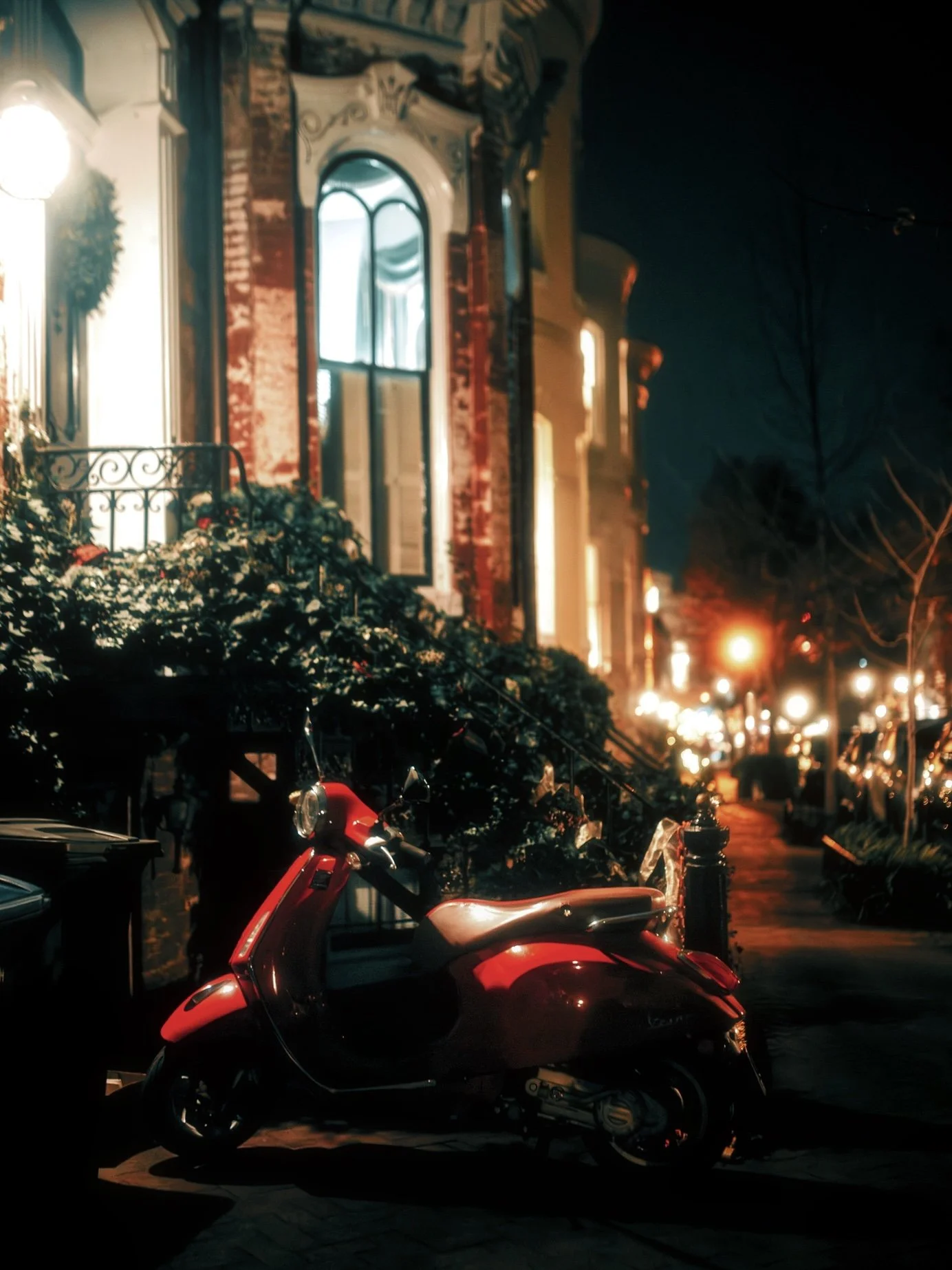 Nighttime street scene featuring a red scooter parked on the sidewalk in front of old-style buildings with brightly lit windows and decorative facades, and streetlights illuminating the street.