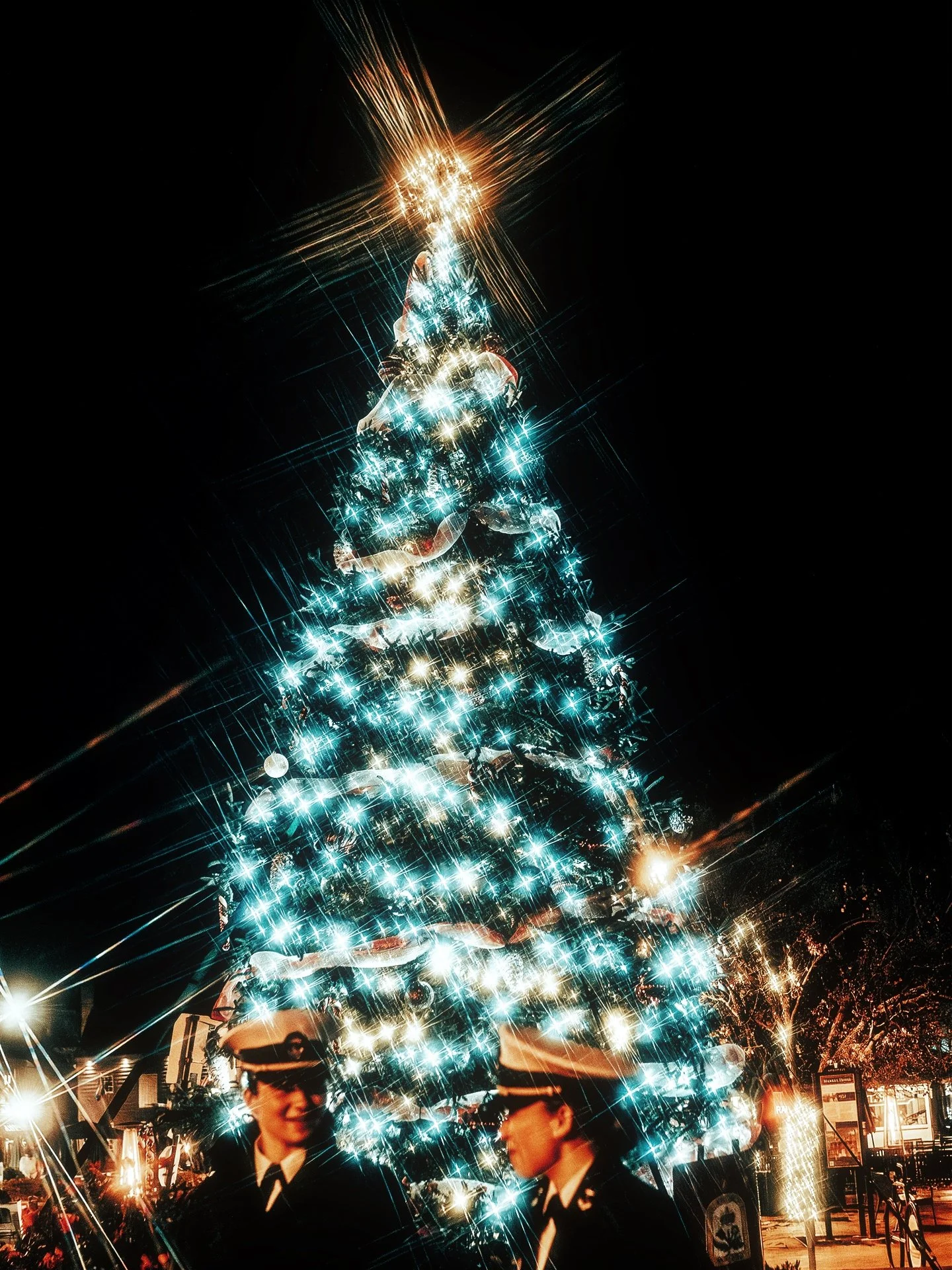 Christmas tree decorated with lights, topped with a star, with two uniformed individuals in front of it at night.
