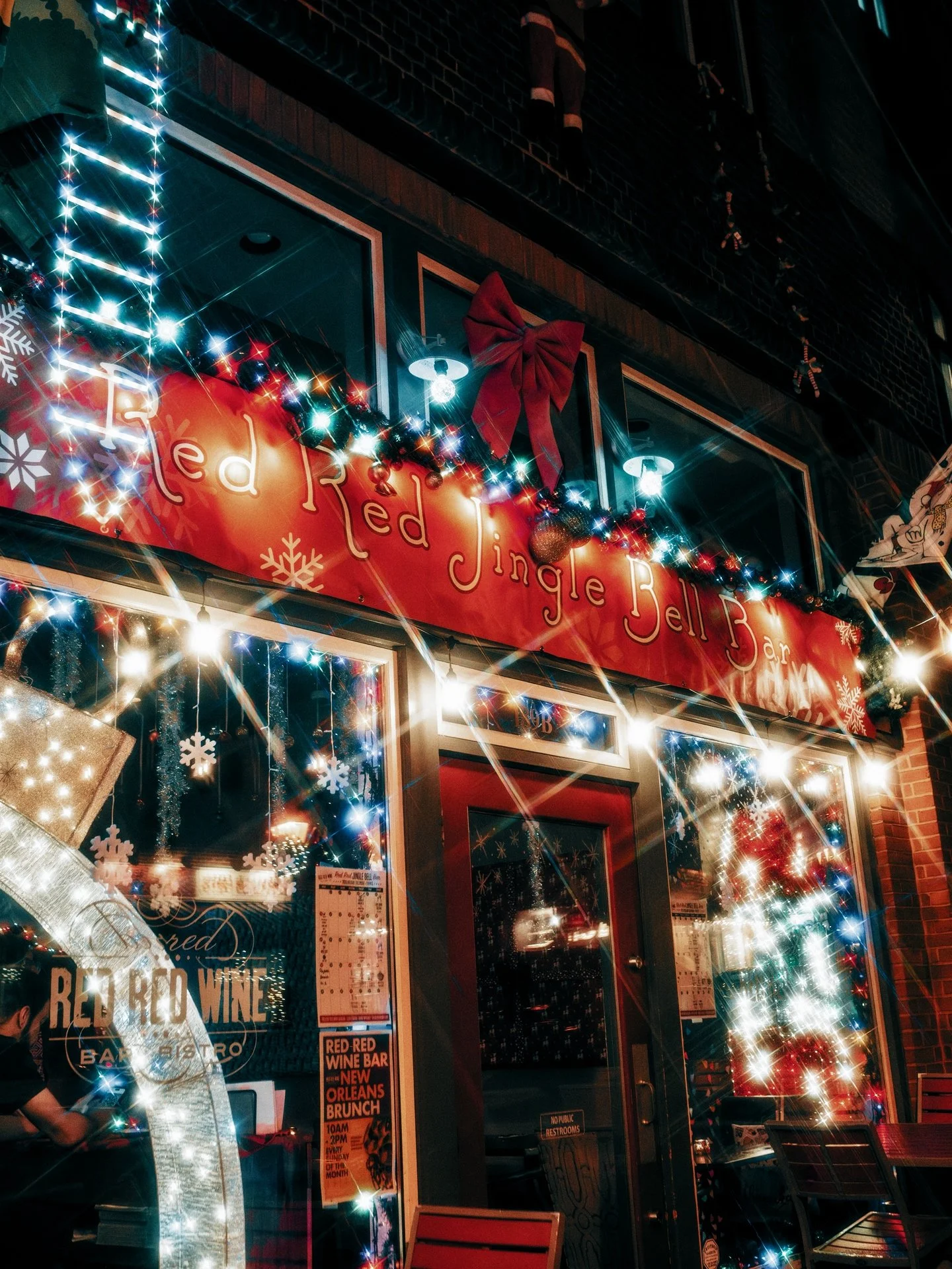 Christmas decorated storefront with red banner reading 'Red Jingle Bell Bar,' adorned with colorful lights, a big red bow, snowflake ornaments, and a Christmas tree visible inside.