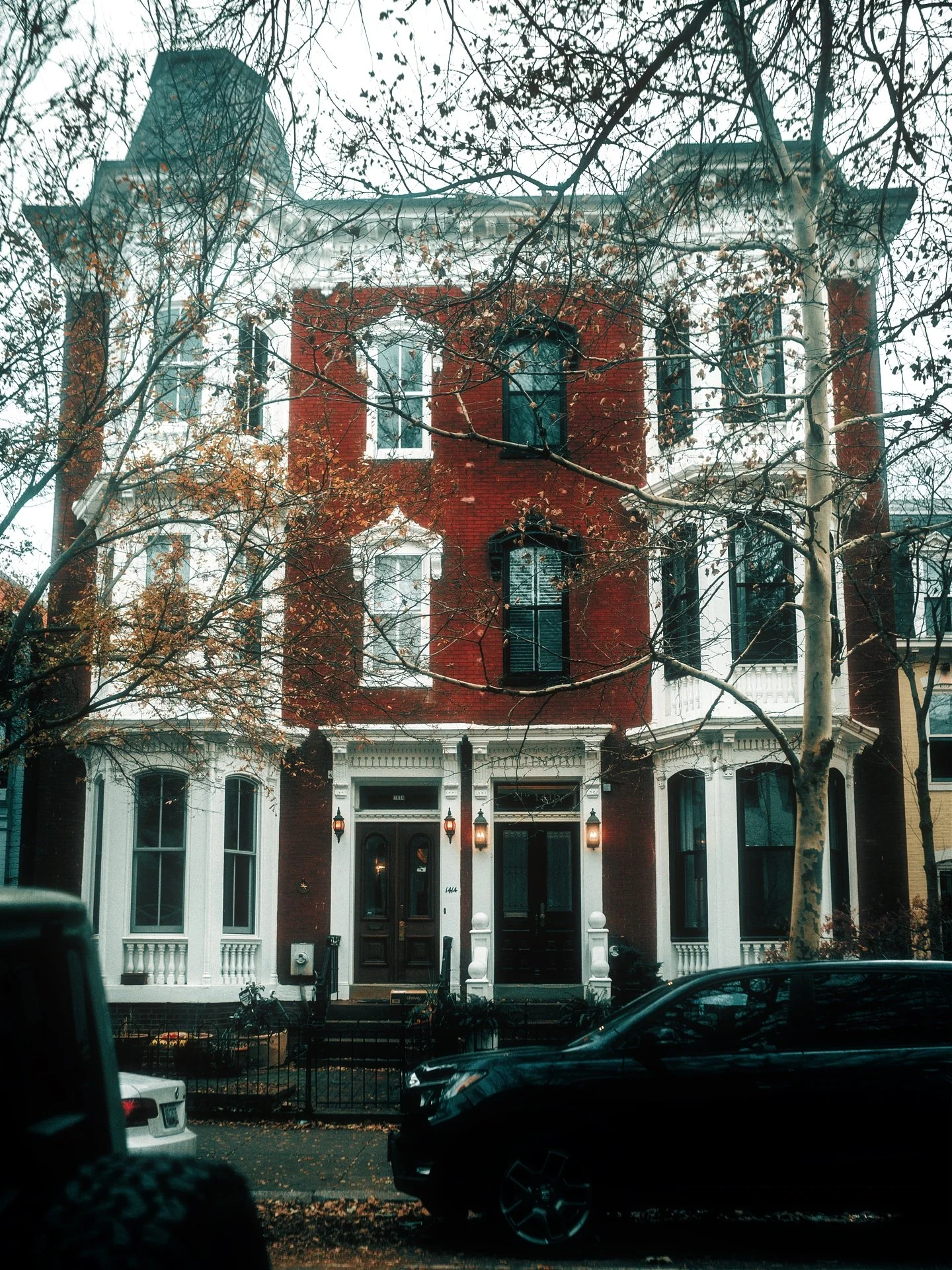 A multi-story row house with a red brick facade, white trim, and large front windows, situated on a landscaped street with leafless trees and parked cars.