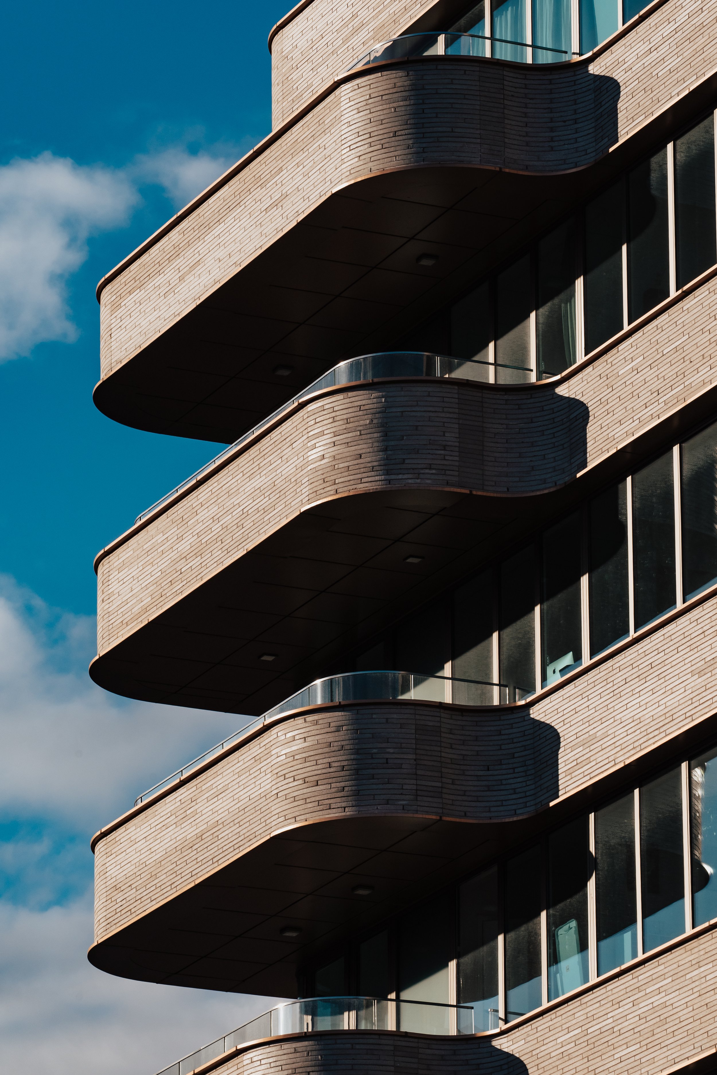 Close-up of a modern apartment building's curved balconies with glass railings, brick exterior, and shadows cast by the structure, against a blue sky with some clouds.