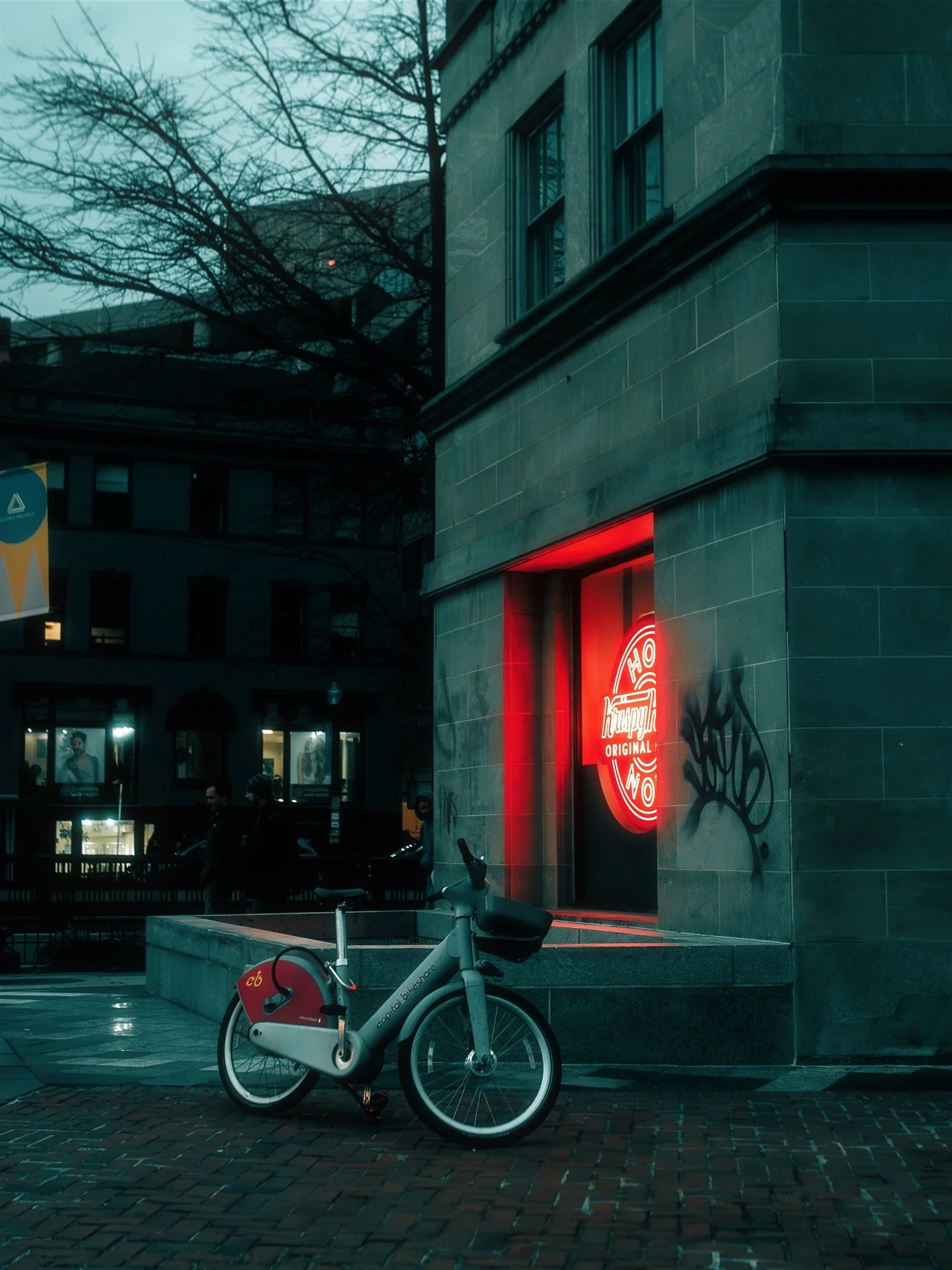 A city street corner with a gray bike parked in front of a building with a red neon sign that reads 'Krazy Wok'. The building has gray stone facade and graffiti on the wall. In the background, there is another building with large windows and some ped