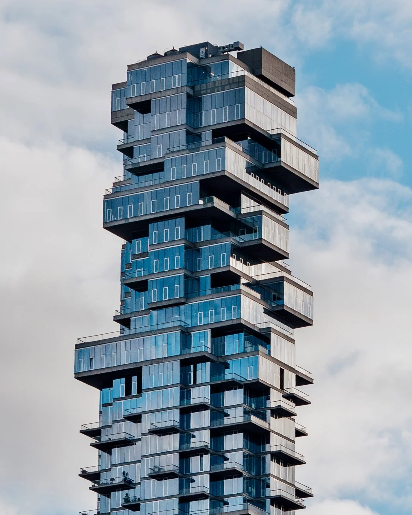 Modern high-rise building with a unique stacked and protruding architectural design, featuring glass windows and balconies under a partly cloudy sky.