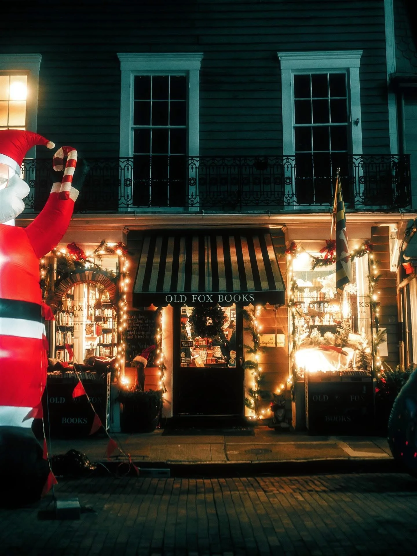 A storefront decorated for Christmas, with string lights, a wreath, and holiday decorations. A large inflatable Santa Claus is partly visible on the left side of the image. The store sign reads 'OLD FOX BOOKS'.