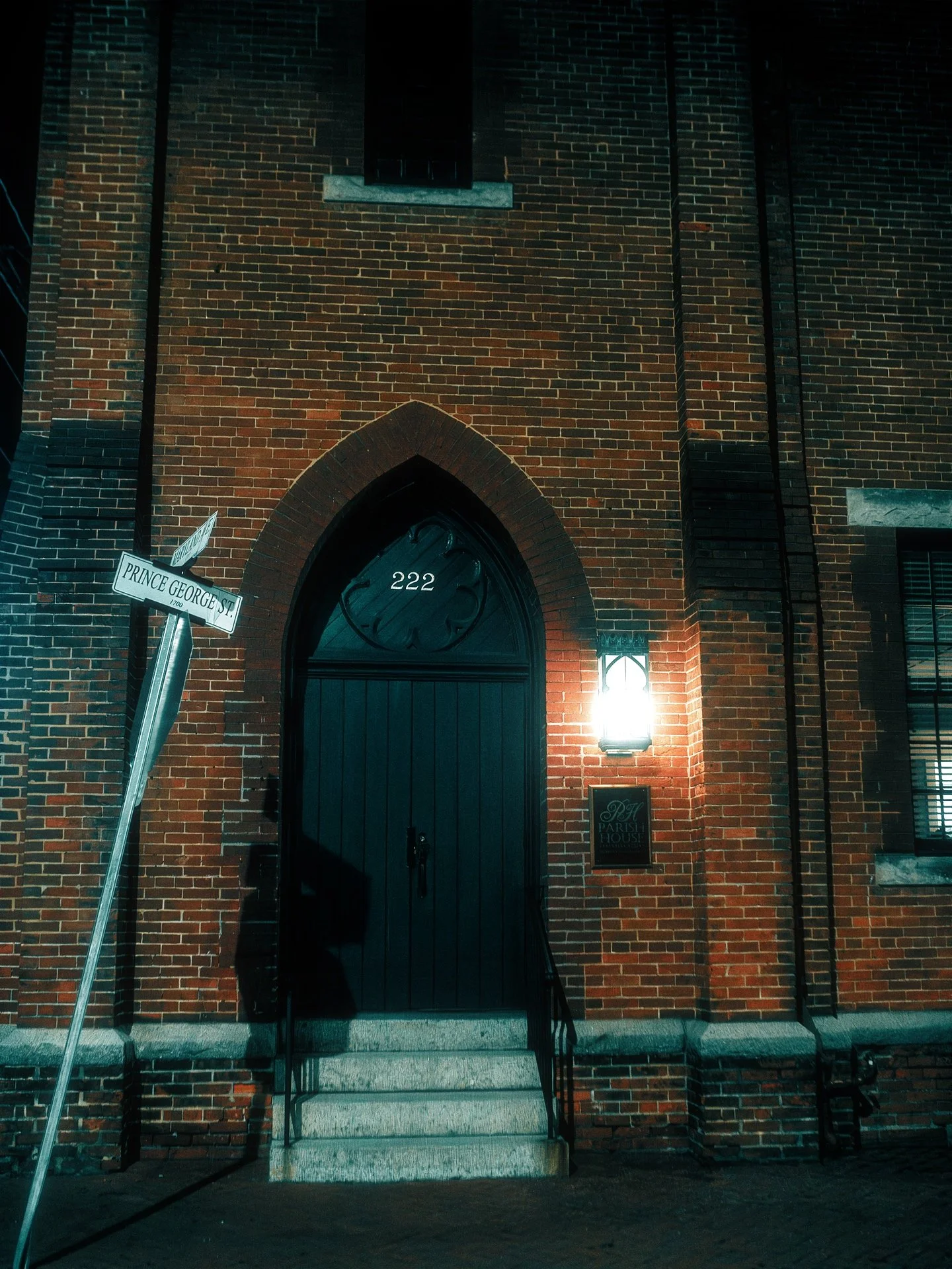 Nighttime view of a brick church building with a black arched door labeled 222, illuminated by a wall-mounted light. A street sign indicates the corner of Prince George Street. The building has a small window above the door and another to the right.