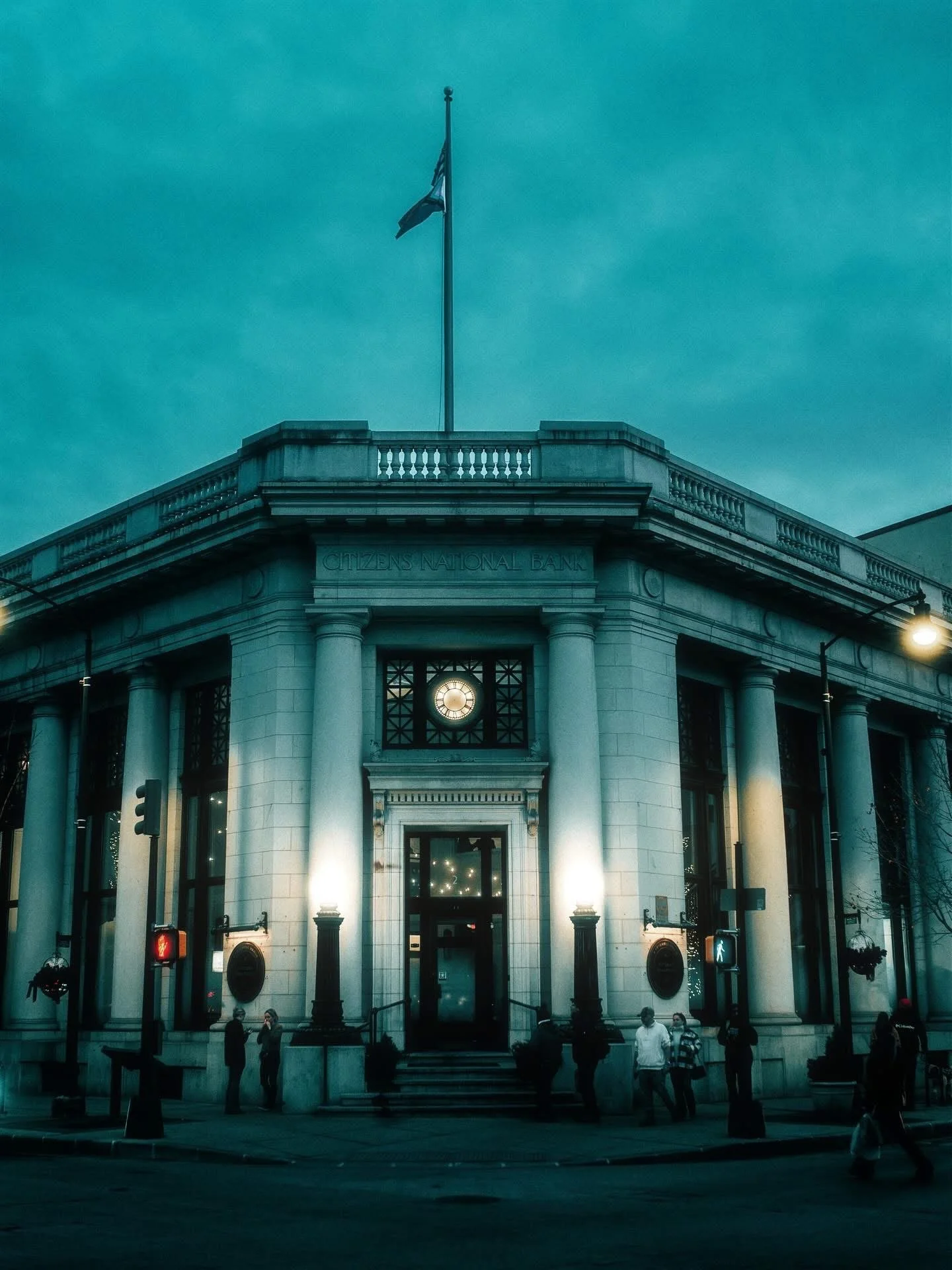 An old bank building with the words 'Citizens National Bank' inscribed above the entrance, a clock above the door, flagpoles with flags on top, and people standing outside at night.