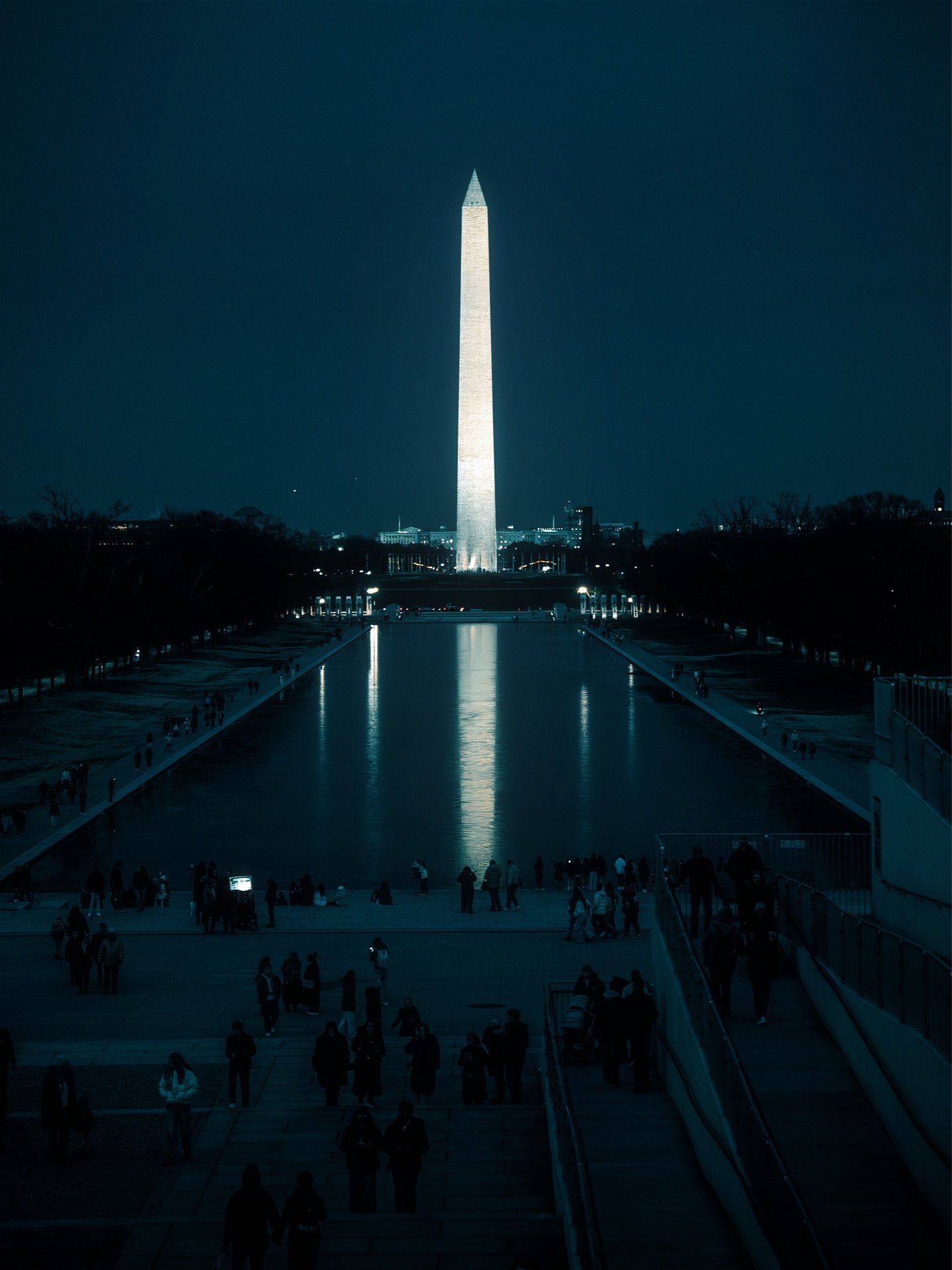 Nighttime view of the Washington Monument reflected in the Reflecting Pool, with people gathered around.