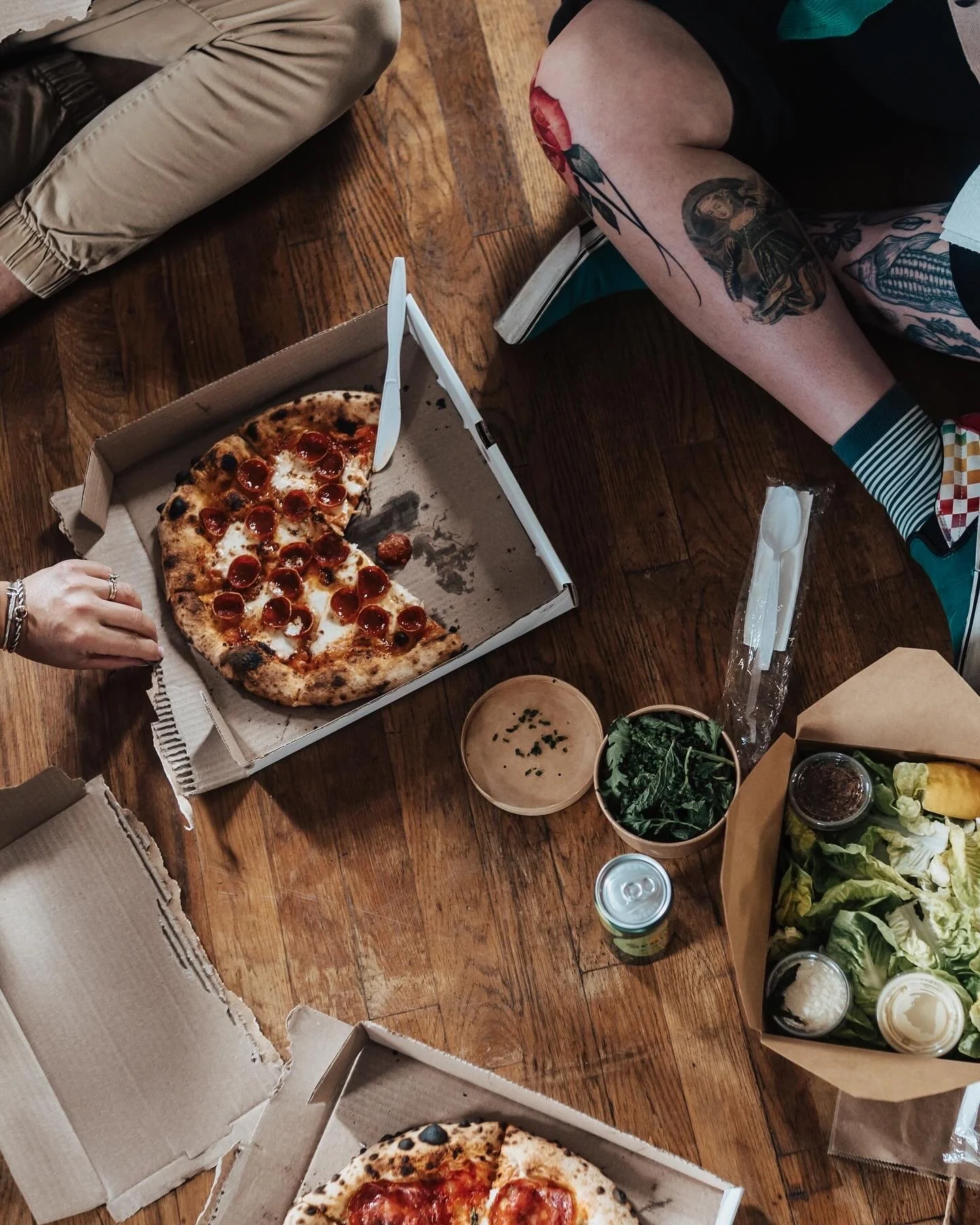 Pizza boxes with partially eaten pepperoni pizza, a salad in a brown paper bag, and drinks on a wooden table, with two people sitting nearby, one with tattoos on their legs.