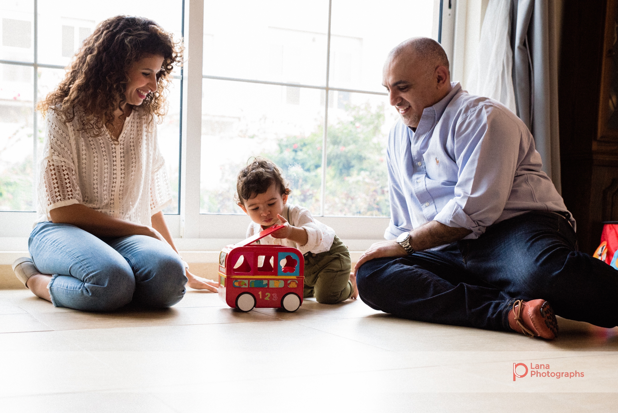 mother and father sit with their one year old baby boy next to the window and play with his toy truck