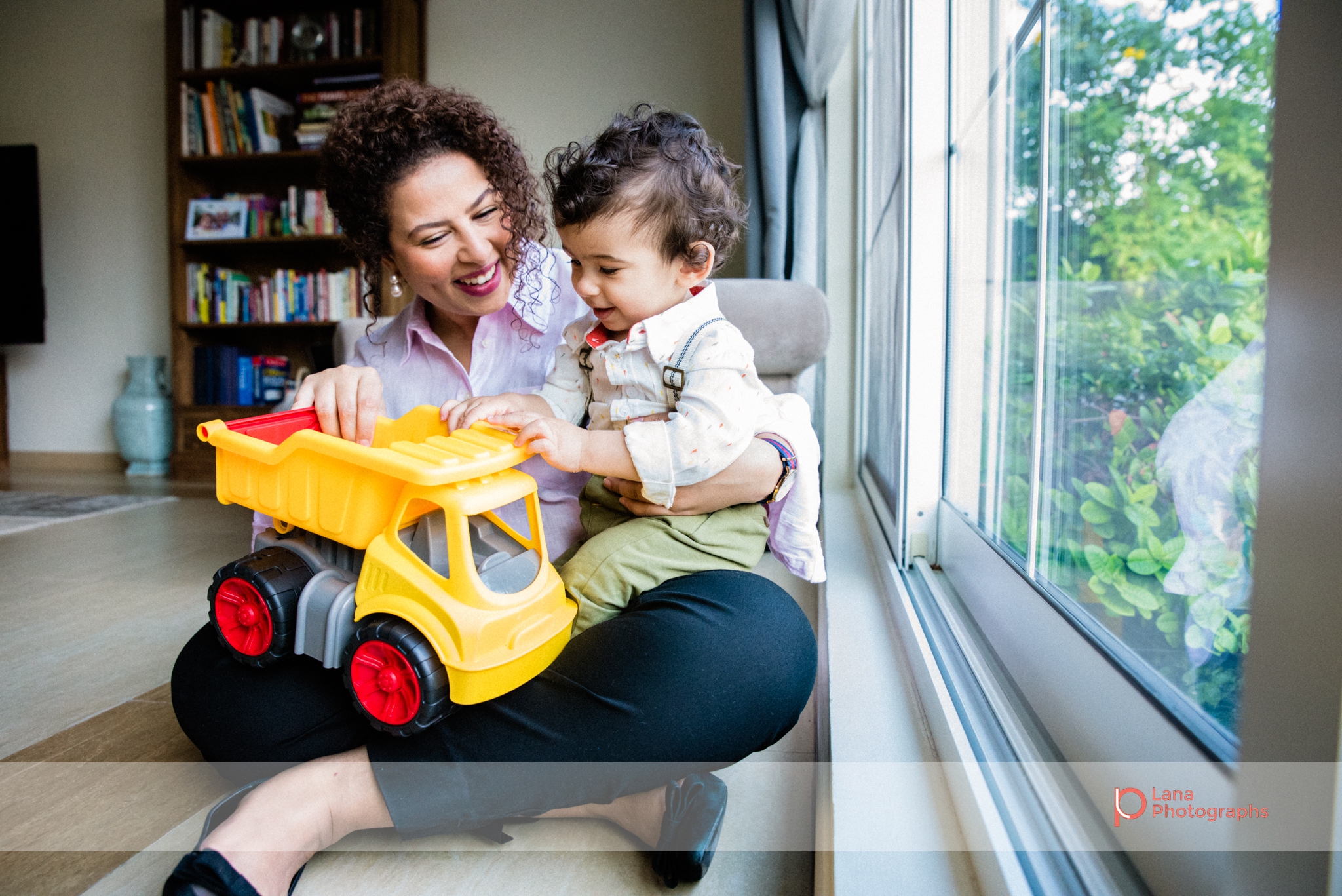 baby and his auntie sit next to the window and play with his yellow toy truck