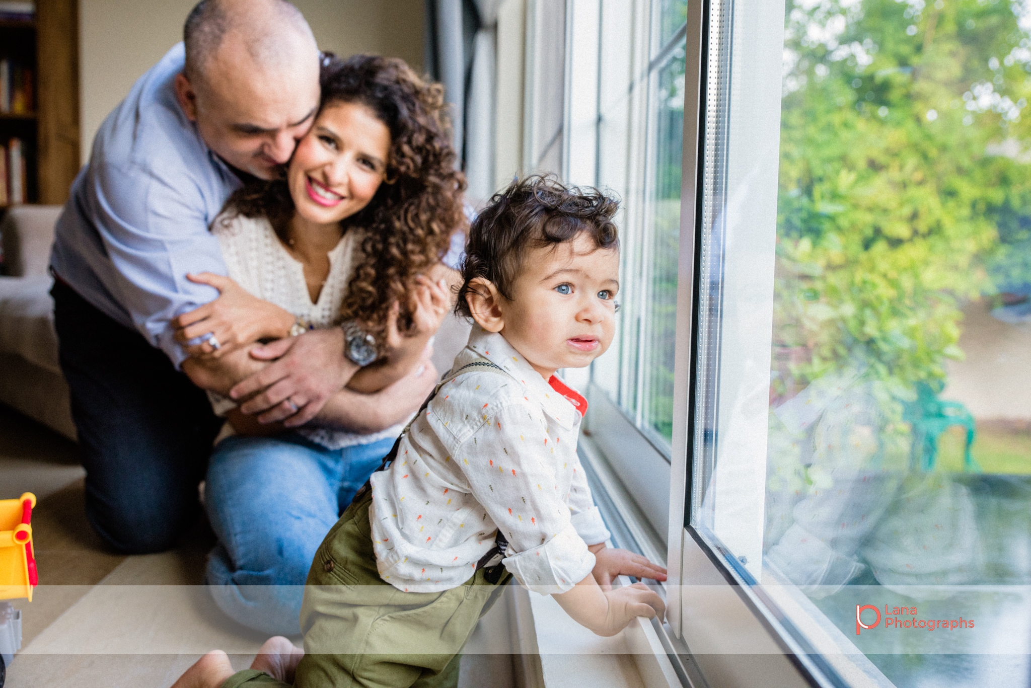 mother and father embrace next to the window in beautiful morning light as baby boy looks outside into the garden