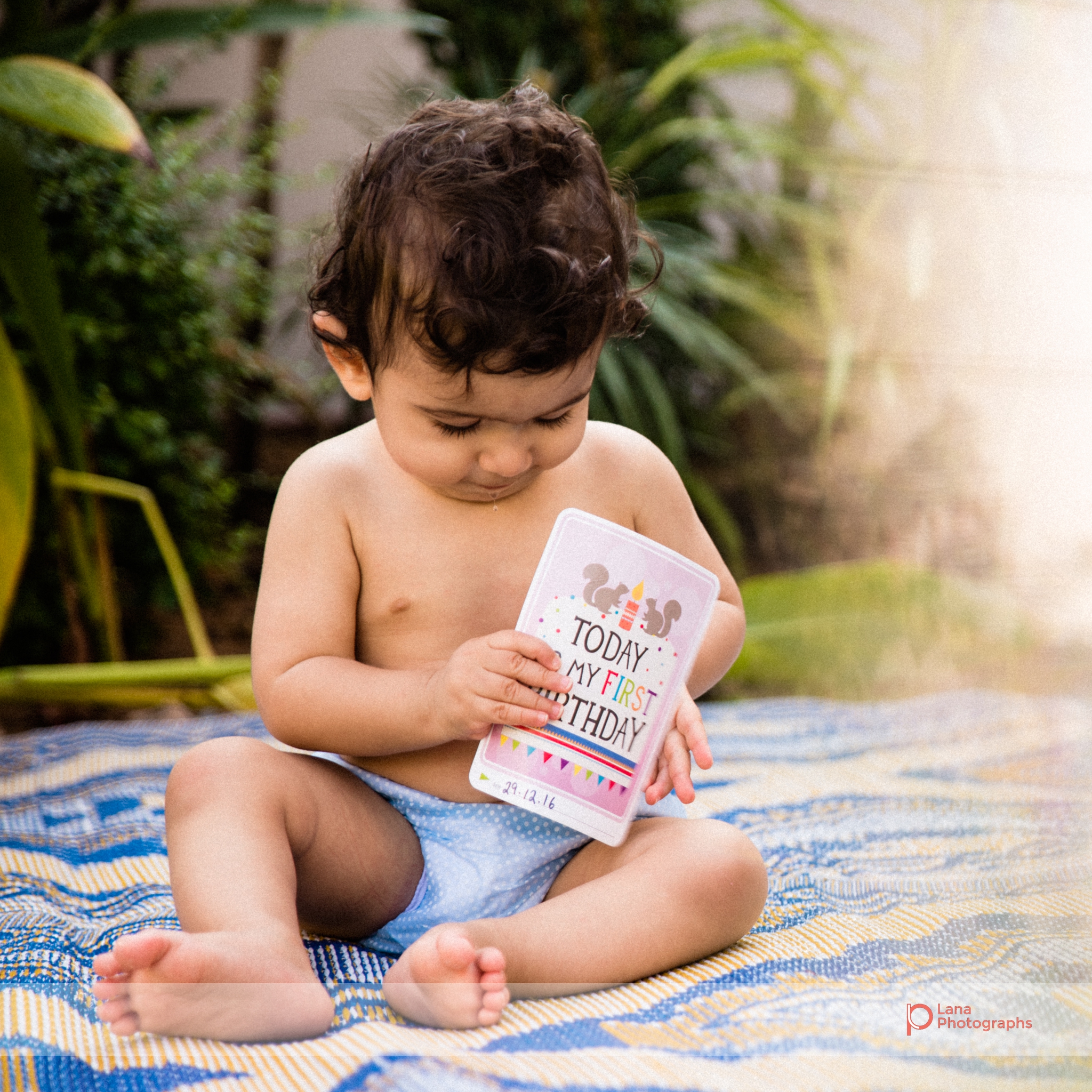 baby boy holds a birthday card and sits on the grass outside his home