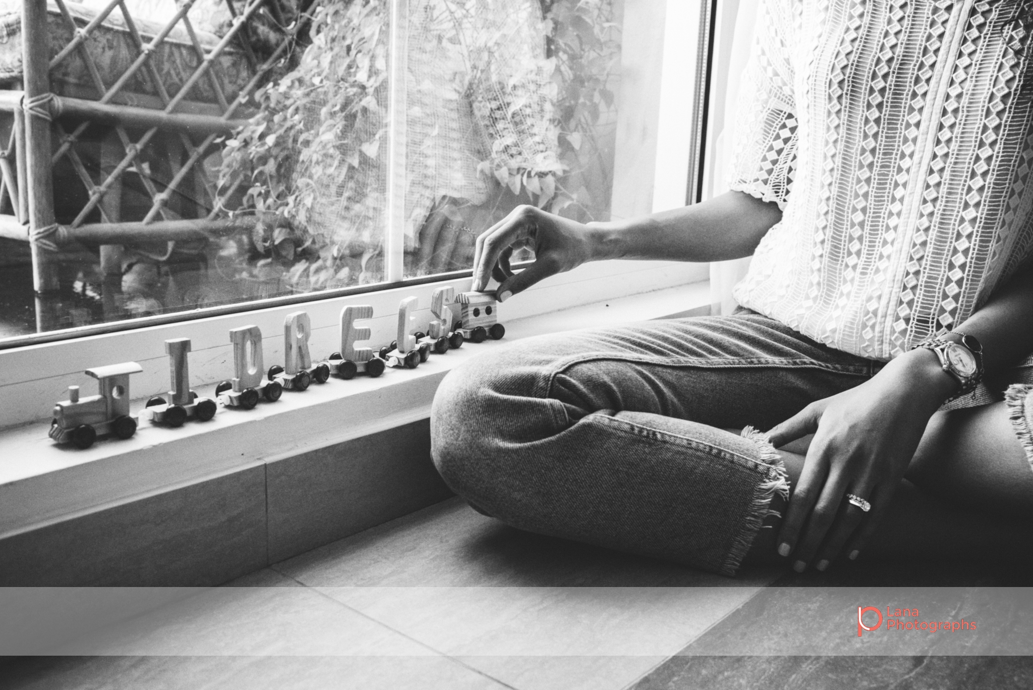 mother sits by window sill in beautiful morning light and plays with wooden train that spells her baby's name