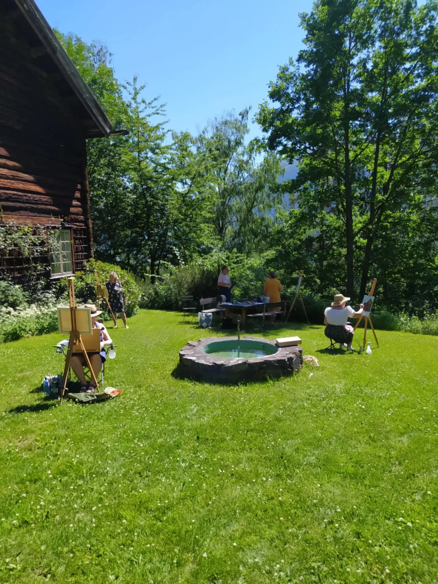 People painting outdoors in a lush green backyard with trees and a small fountain, under a clear blue sky. Painting course at Nutheim