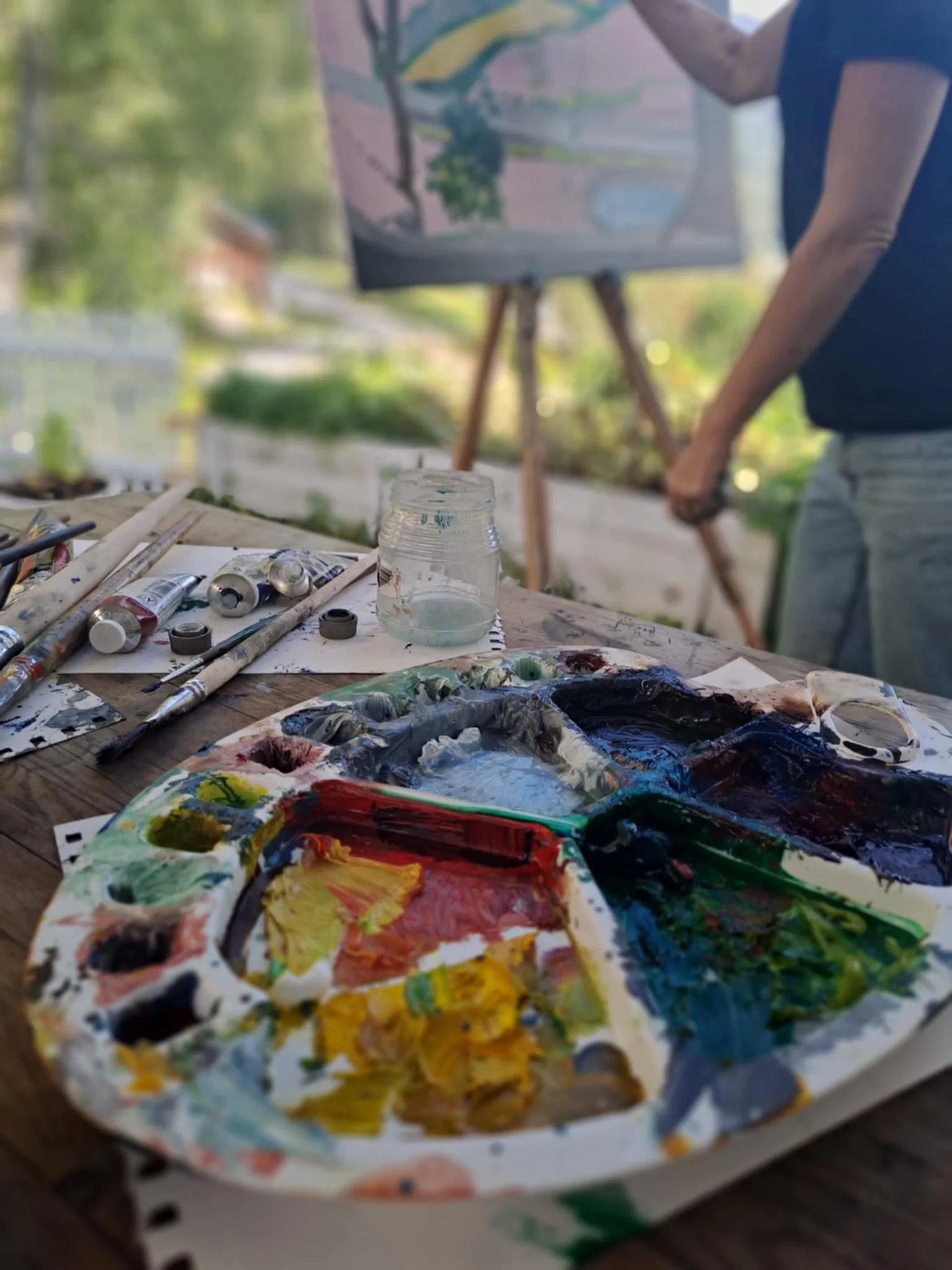 Paint palette with mixed colors, paintbrushes, and art supplies on a wooden table outdoors with a person painting on an easel in the background.