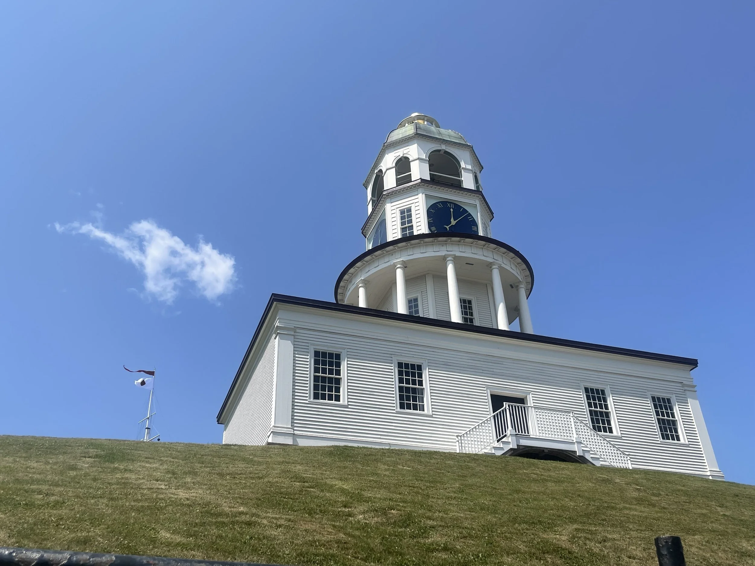 A white historic building on a green grassy hill, blue sky in the background with one lone cloud.
