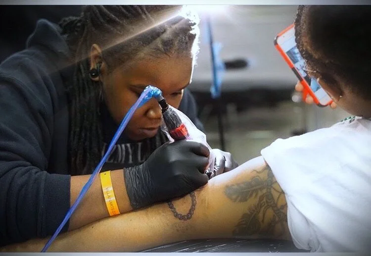 A woman is getting a tattoo tattooed on her forearm by an artist wearing gloves, while another person looks on with a phone recording.