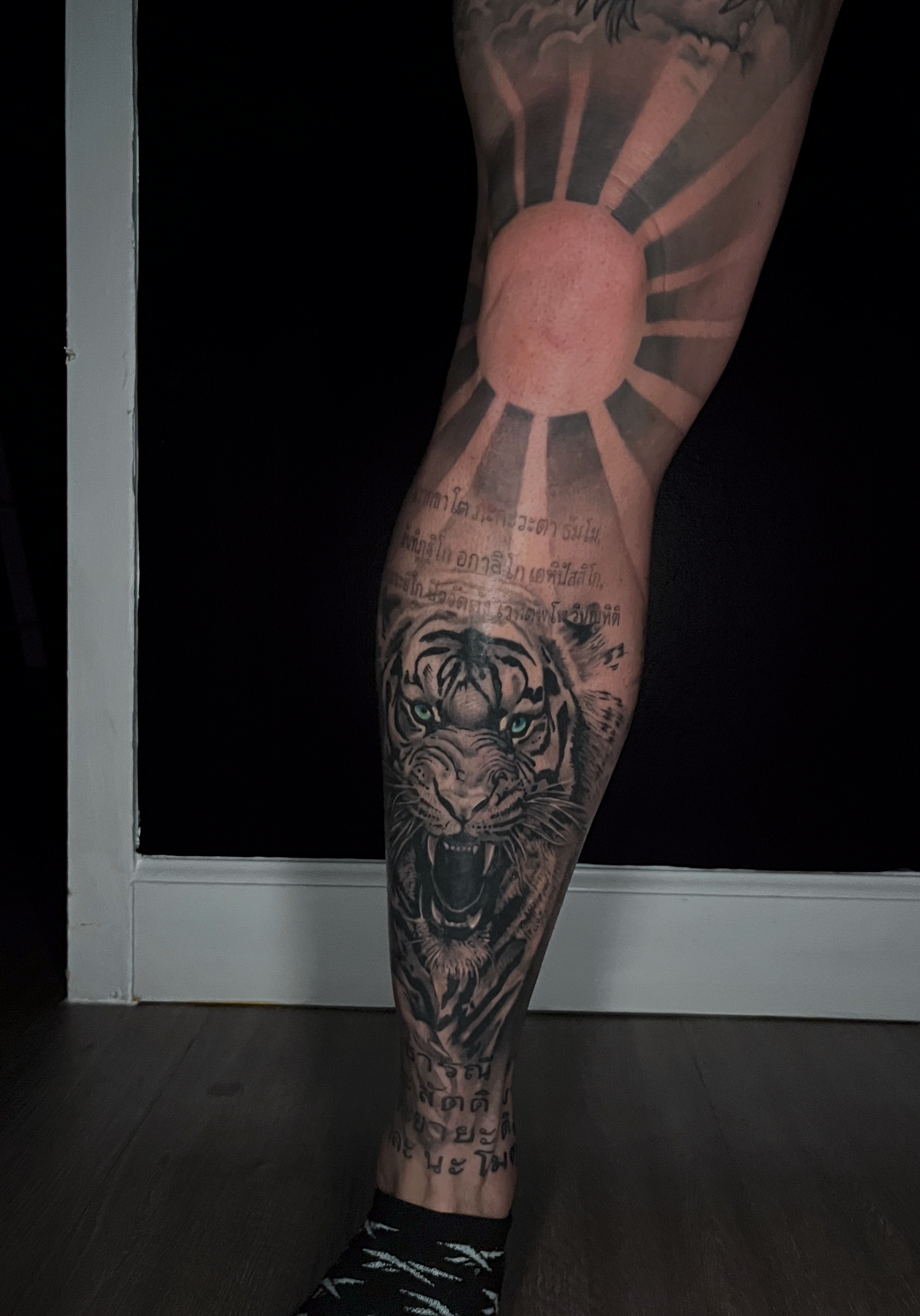 Tattoo of a roaring tiger with glowing eyes on a person's lower leg, with some Hindi or Thai script above and below the tiger, and a large sun or celestial body with rays behind the leg.