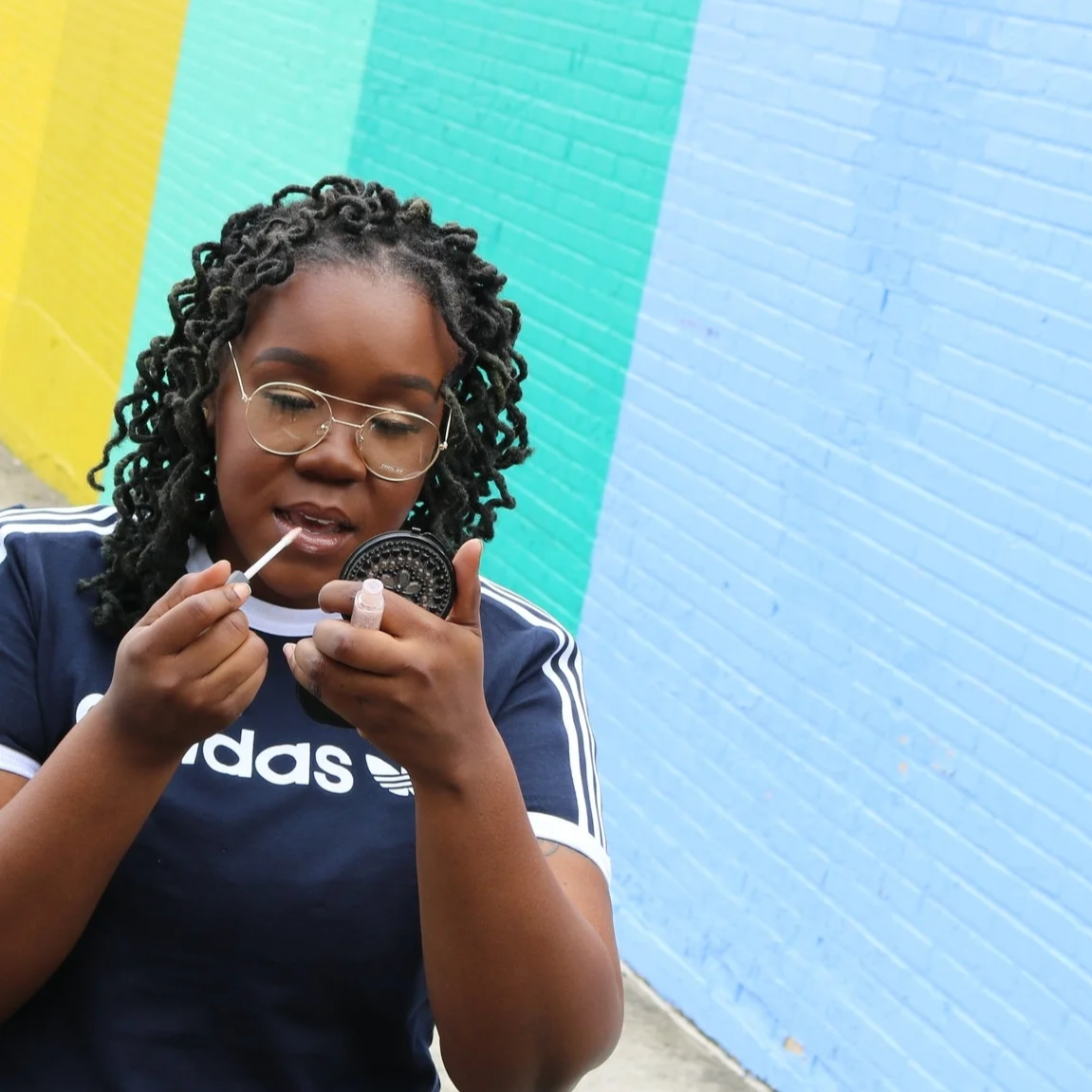 Young woman with dreadlocks and glasses applying lip gloss or lipstick outdoors against a colorful mural wall.