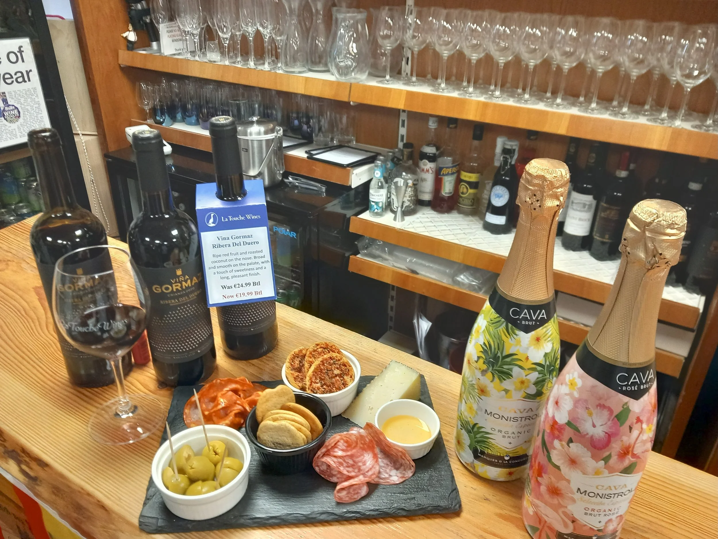 A wooden bar counter with two bottles of sparkling wine, a glass of red wine, and a plate of assorted charcuterie, cheeses, olives, and crackers. The background features shelves of glassware and liquor bottles.