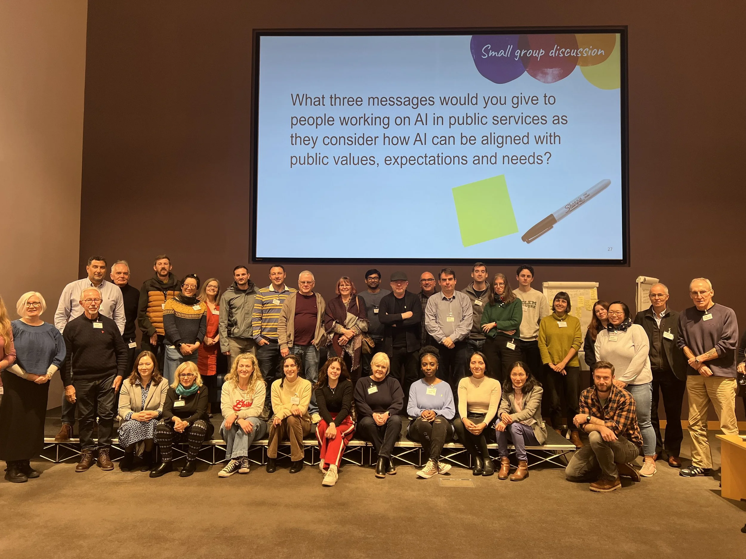 A diverse group of people sitting and standing in front of a large screen which says 'what 3 messages would you give to people working on AI in public services'