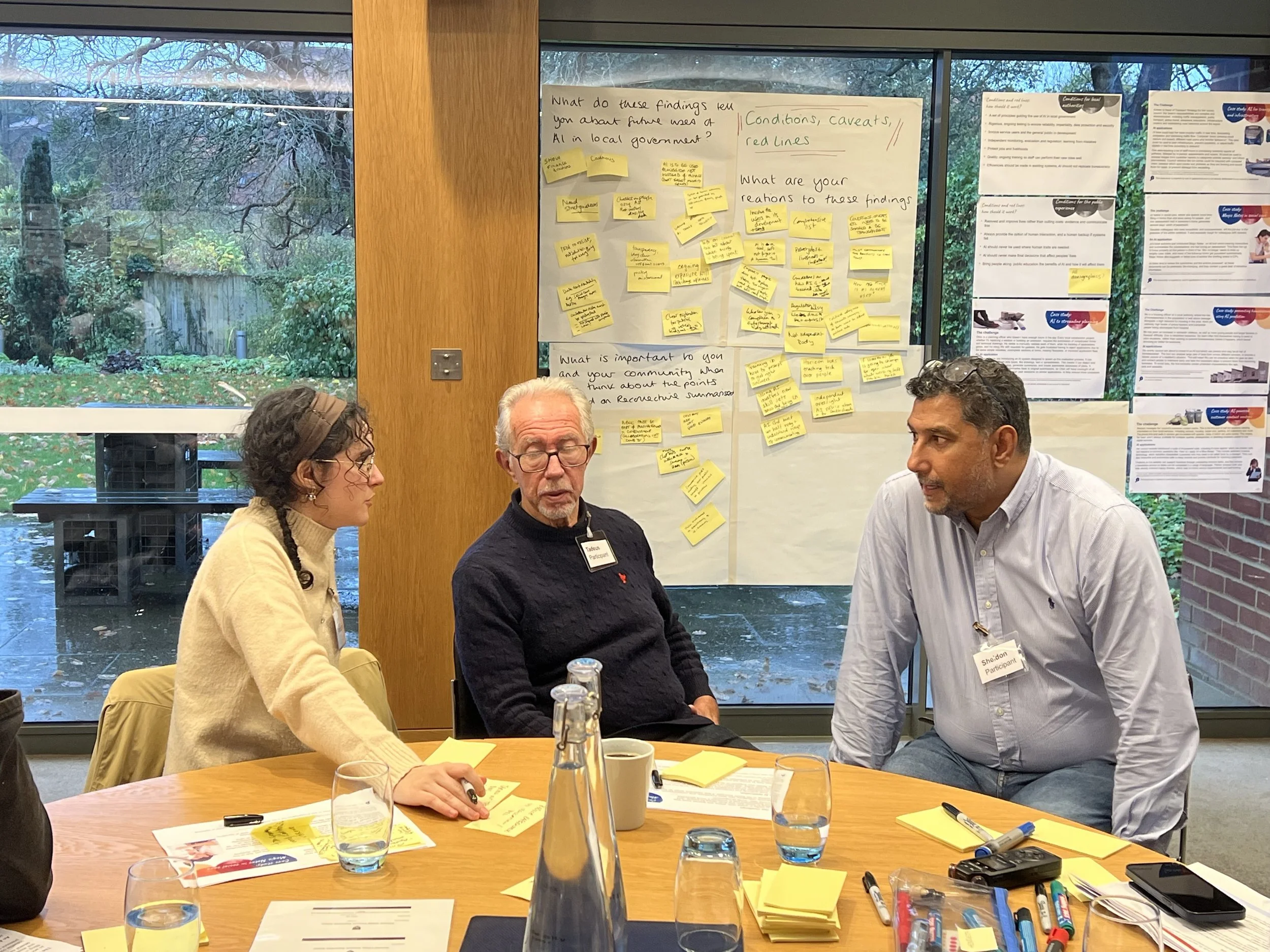 Three people sitting at a table in a room with flip charts and pens around them. They are discussing AI in local government.