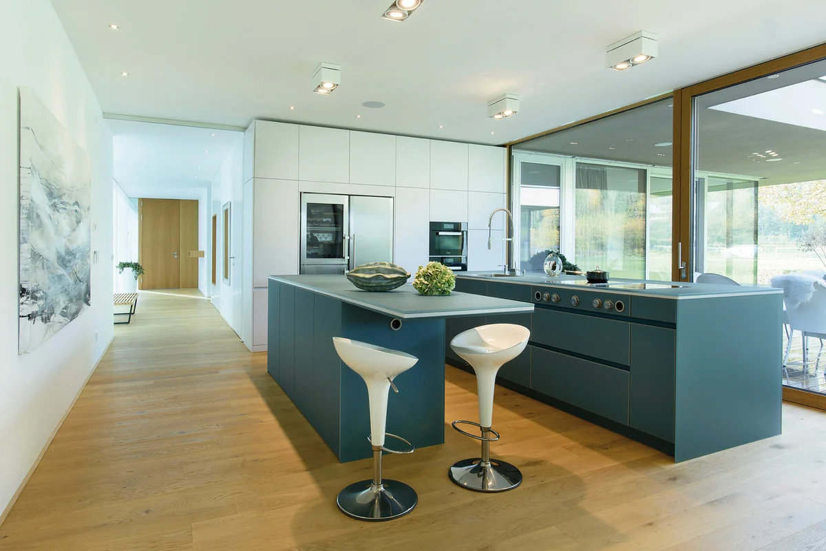 The house is flooded with light and the floor-to-ceiling windows open out over a vista of the meadow with an orchard and clumps of trees. The kitchen and living room areas are at an angle to the veranda, giving rise to an intense connection between exterior and interior.
