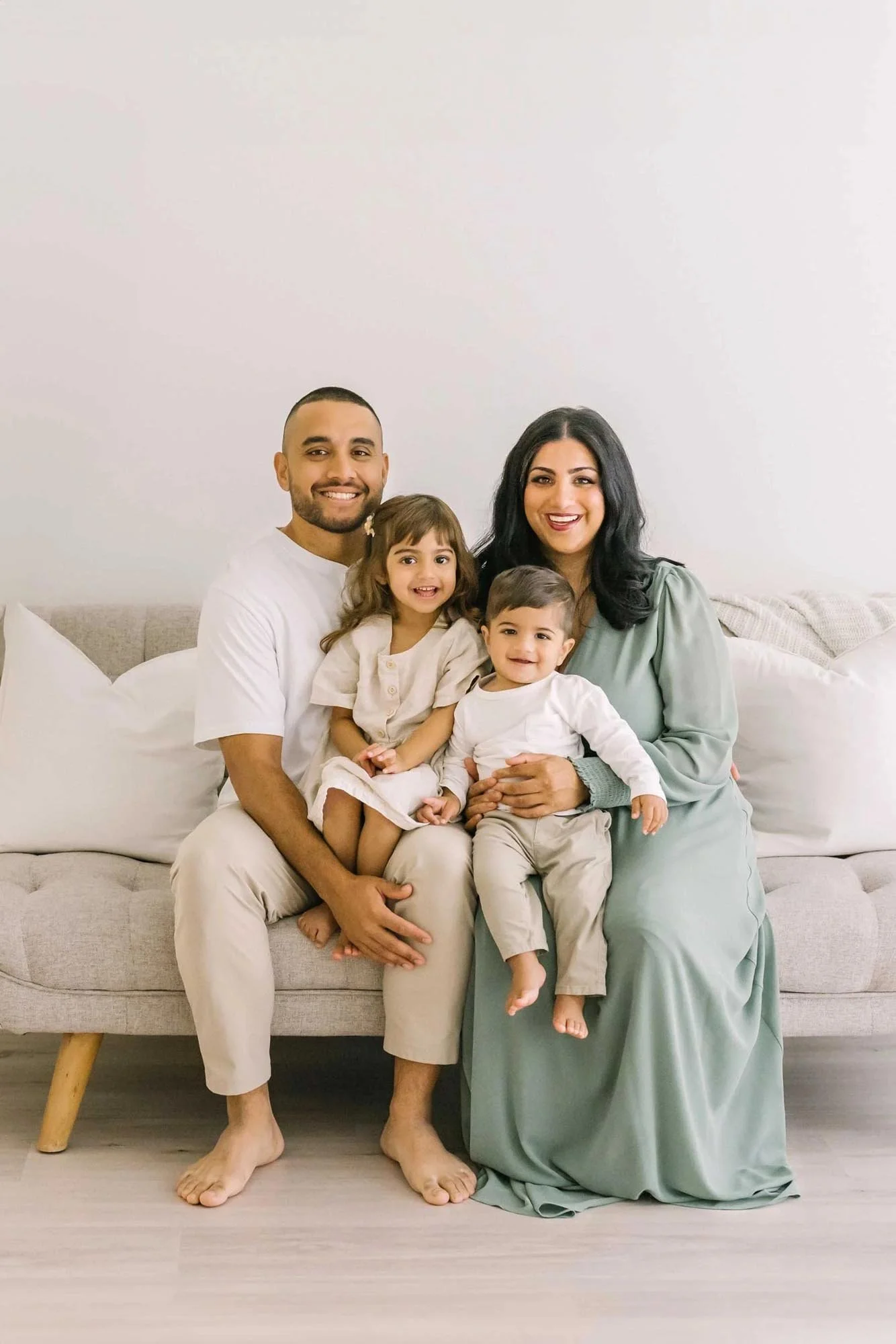 A smiling family sitting on a sofa at family photography studio in Hamilton, Ontario, , with a man, woman, and two young children. The man wears a white shirt and beige pants, the woman in a green dress, and the children in light-colored outfits.