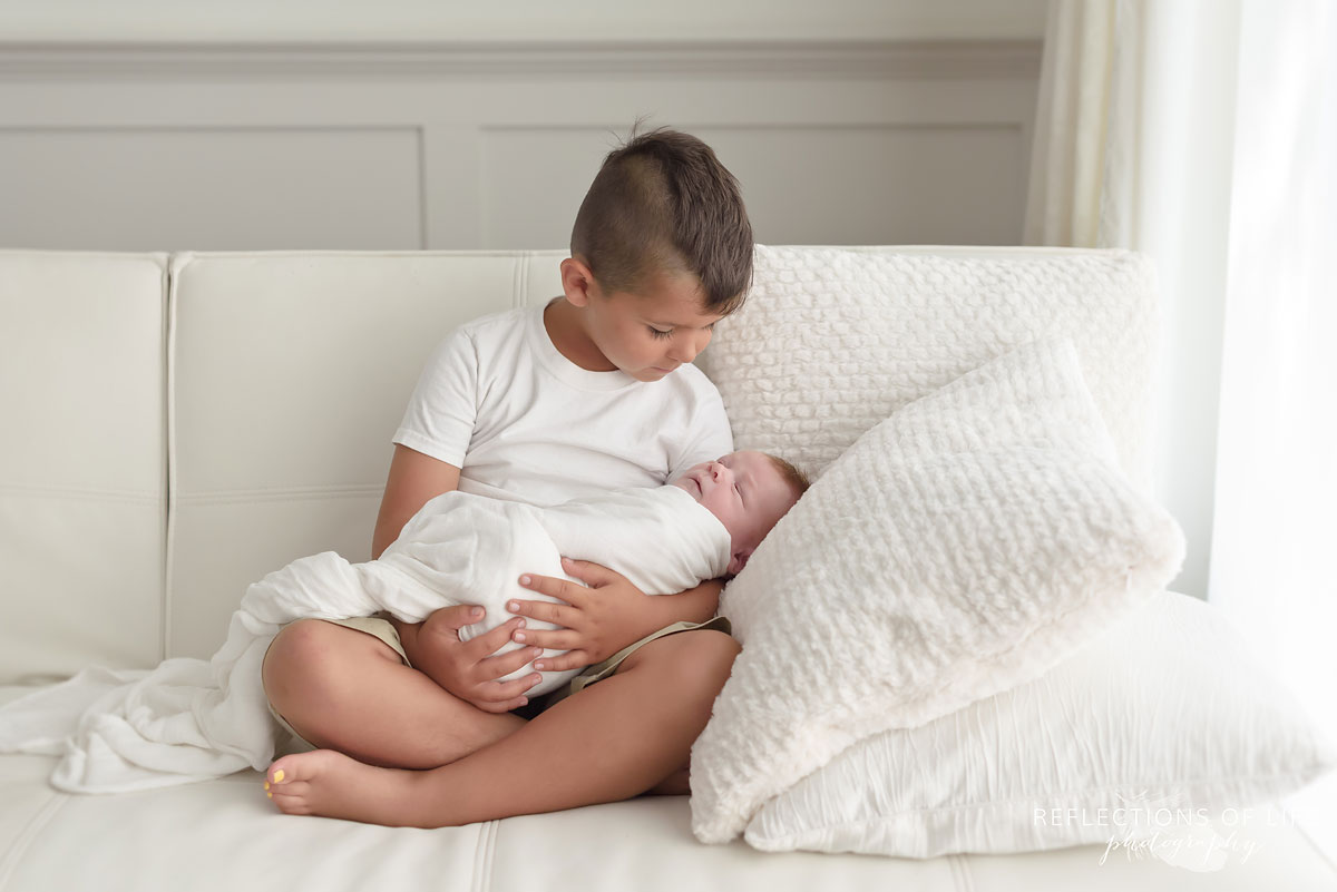 Boy cousin holds newborn baby boy in his arms in white baby photo studio