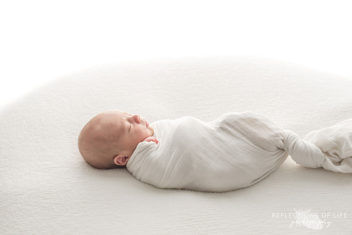 Newborn baby boy swaddled in white on white beanbag backlit with natural light