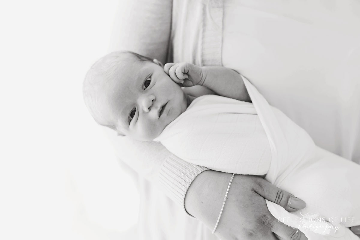 Newborn baby boy looknig at the camera swaddled in white