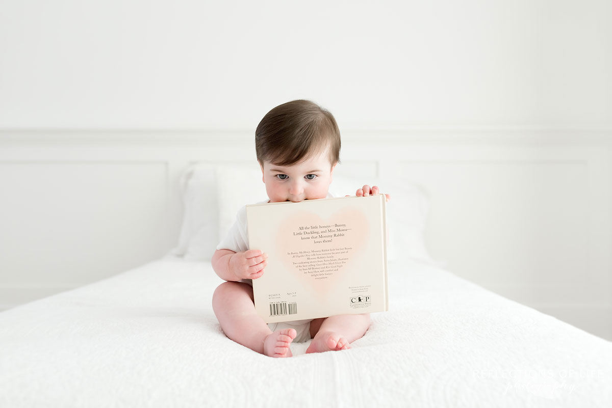 Baby putting edge of a book in mouth on white couch