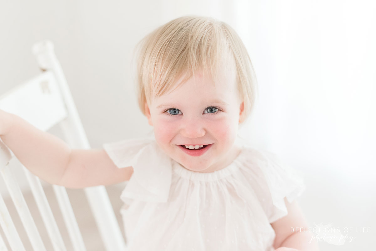 little girl looks totally adorable while sitting on white chair in natural light studio