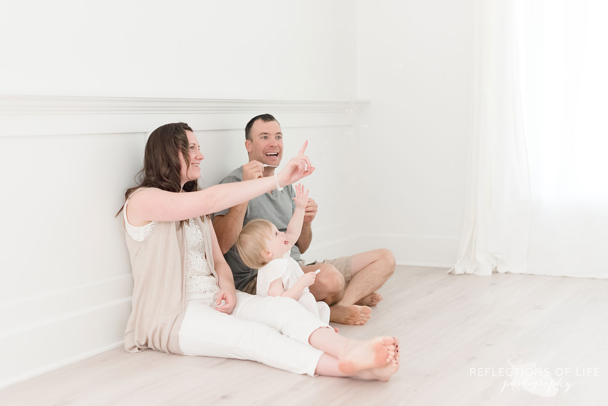 mom and dad blow bubbles with their daughter in natural light studio