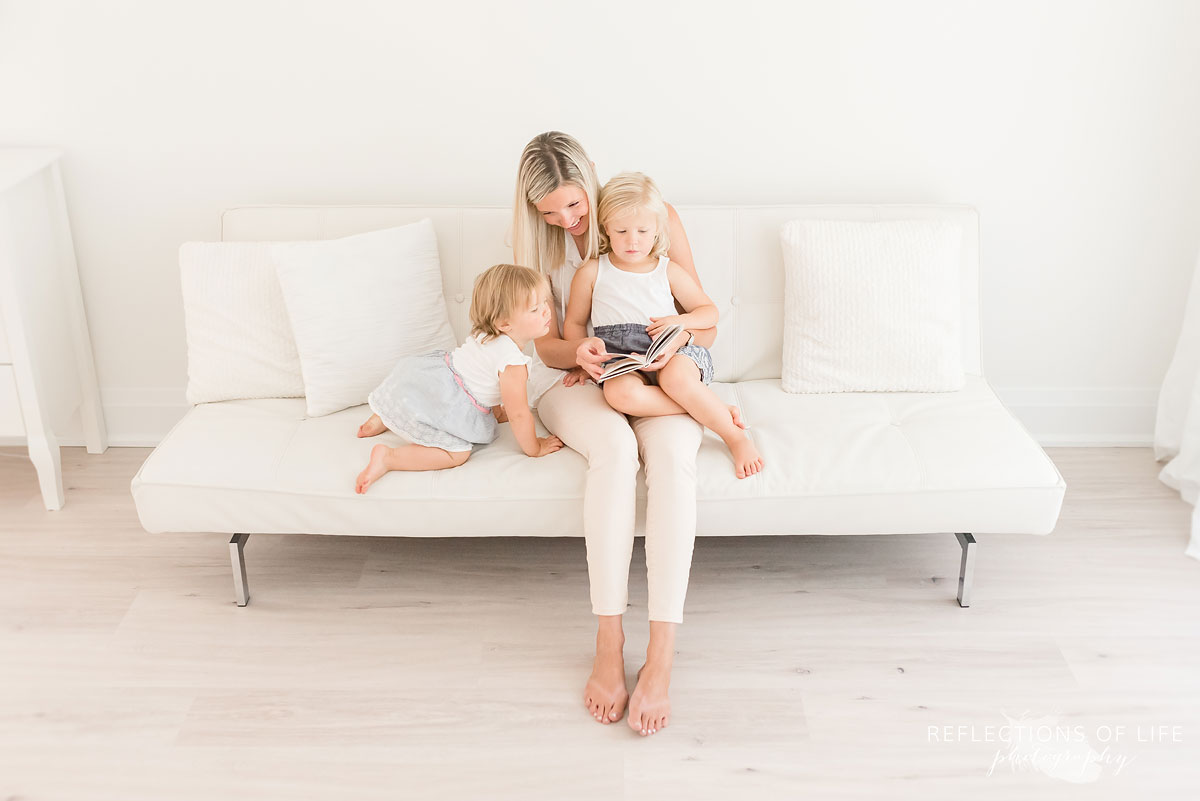 mom and daughters reading a book on white couch