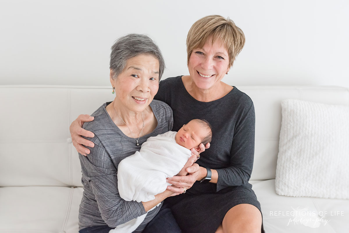 two grandmothers holding their newborn grandchild smiling by Karen Byker