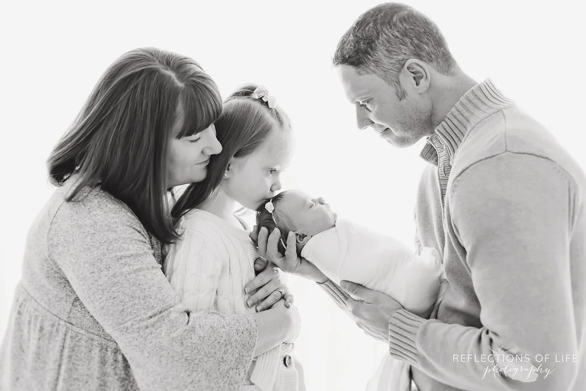 Family of four observes baby while daughter kisses their head black and white.jpg